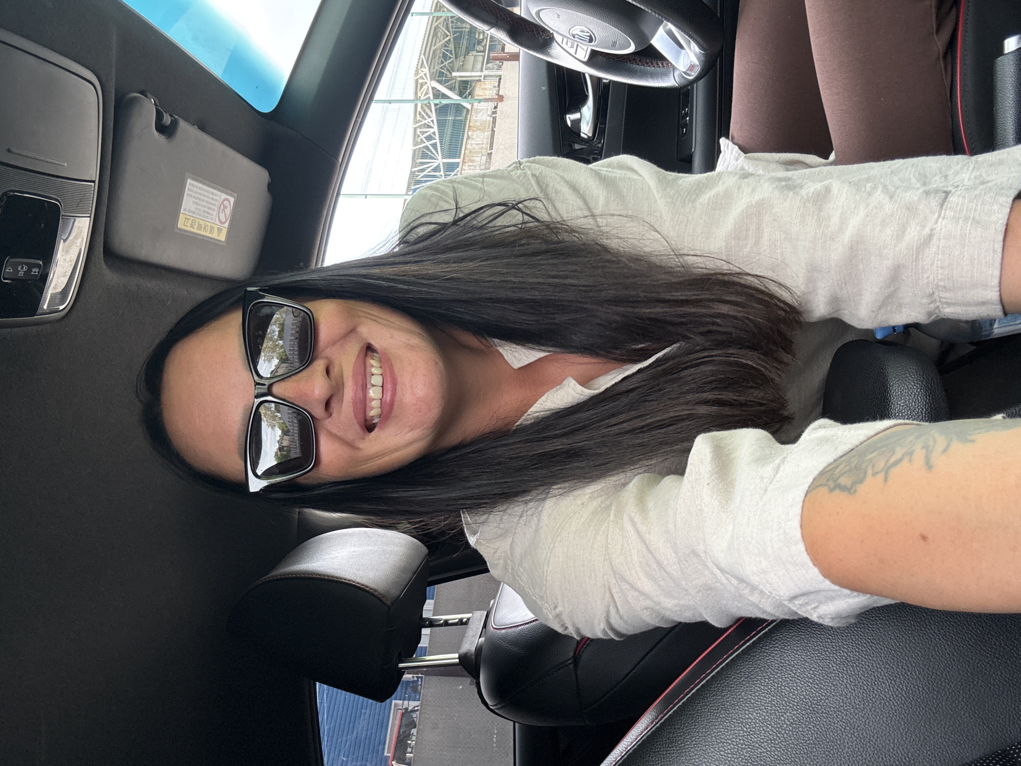 A woman is pictured inside a car, sitting in the driver's seat and looking at the camera. She is wearing sunglasses and a light-colored shirt. Her dark hair is long and falls past her shoulders. Her arms and legs are visible, and she has a tattoo on her left arm. The car's interior is visible, including the steering wheel, dashboard, and seats. The background outside the car appears to be an outdoor area with some structures and fencing. The image does not provide any information about the specific location within the city of Chaikovsky, Russia.