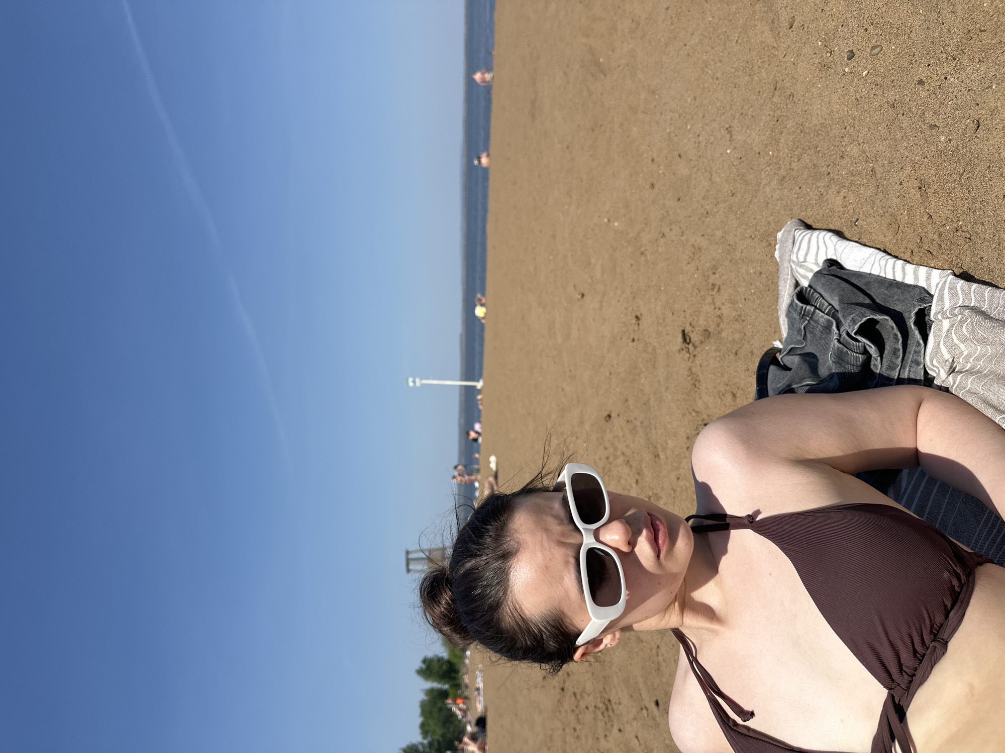 The image depicts a woman in a brown bikini lying on a beach with white sunglasses on. She is on a striped towel with clothes nearby. In the background, there is a beach scene with people, the ocean, and a blue sky with a contrail. The location is Chaikovsky, Russia.