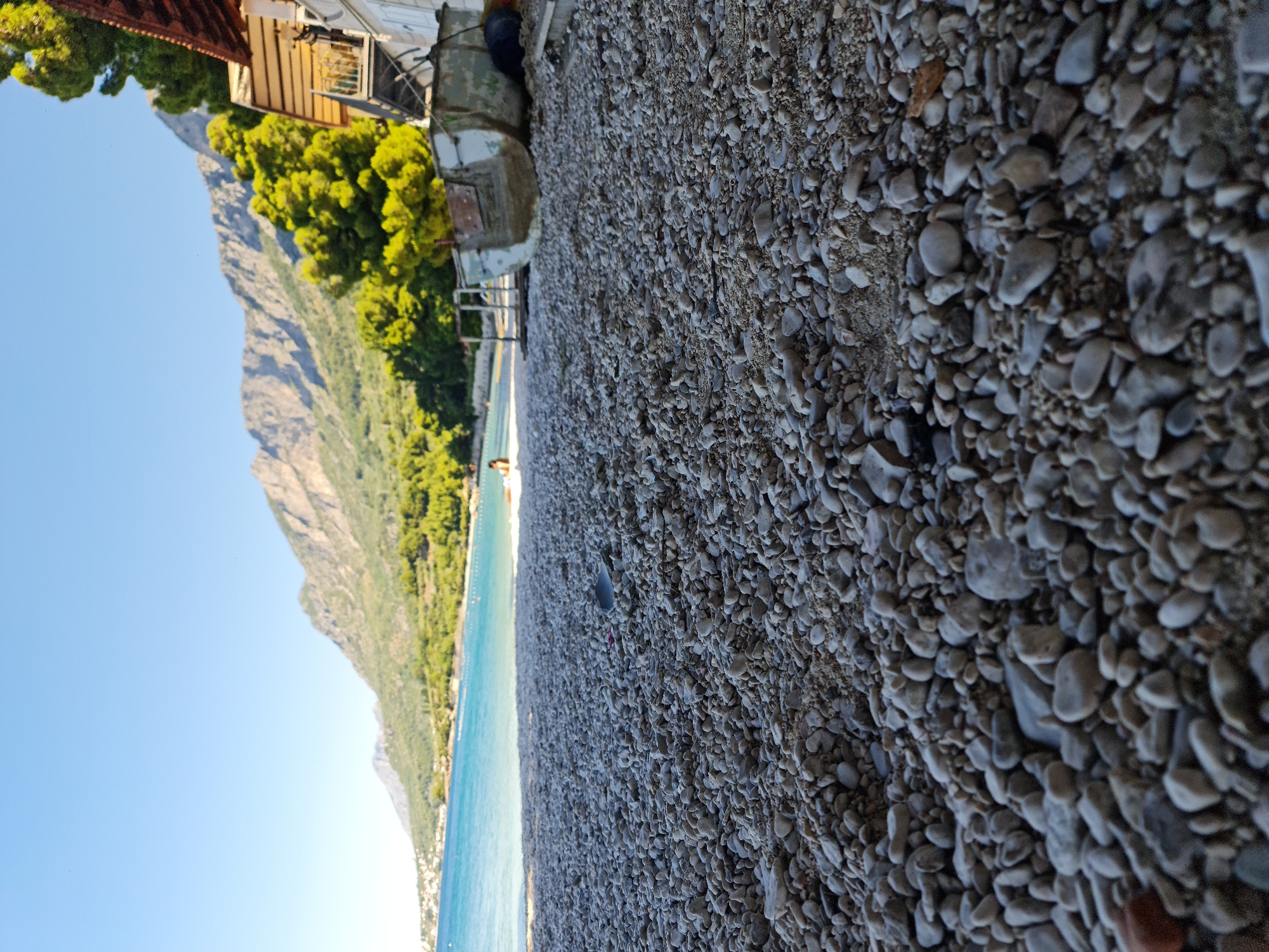 The image depicts a low-angle, wide shot of a pebble beach in Općina Gradac, Croatia. The foreground is dominated by a dense layer of small, grey and white pebbles, with a few larger stones interspersed. A thin strip of turquoise water with gentle waves borders the beach in the mid-ground, with a white buoys in the water. A person is visible as a small silhouette in the distance, standing near the water's edge. Behind the beach, a line of green pine trees meets a steep, rocky mountain range that stretches across the horizon under a clear blue sky. A weathered, partially visible boat, resting on its side, is situated to the right of the frame, near a building with a brown-tiled roof and white walls, suggesting it is a coastal structure or a dwelling. The overall scene conveys a serene, natural coastal environment.