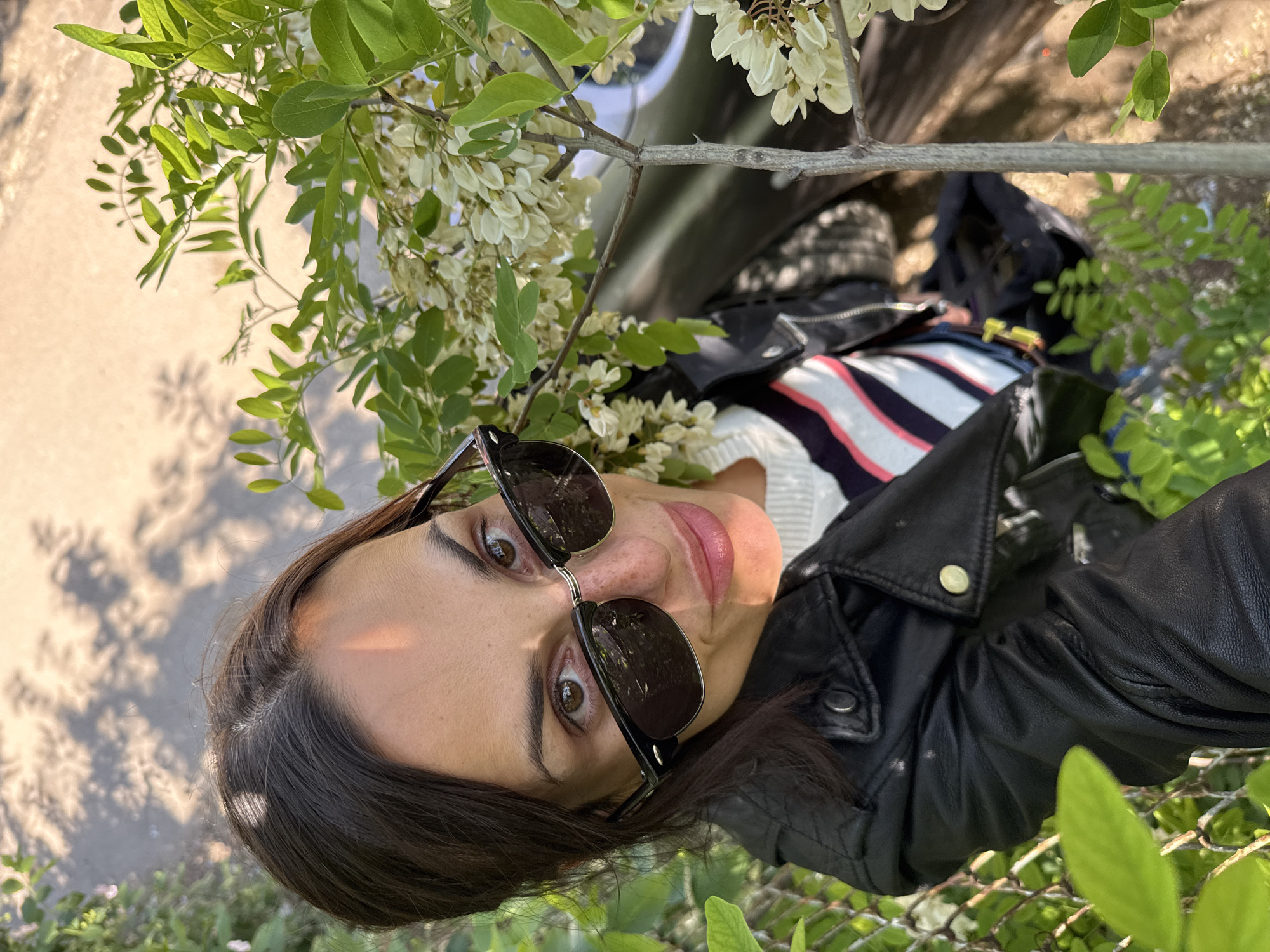 A woman with dark hair and sunglasses is taking a selfie from a low angle. She wears a black leather jacket and a striped shirt. White flowers and green foliage surround her. The image was taken in Pleven, Bulgaria.