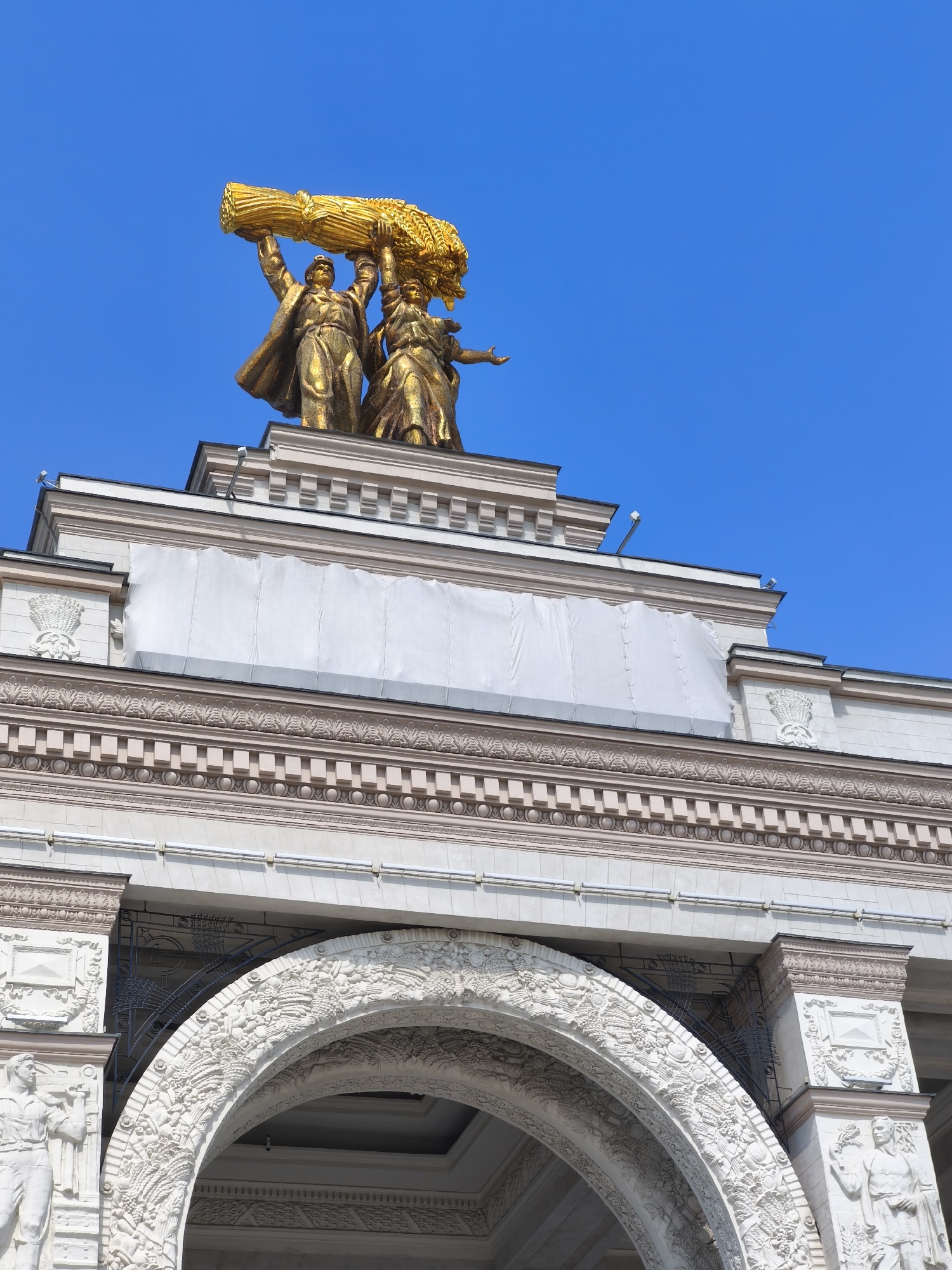 In the city of Unknown, Unknown, a monumental golden statue of a man and a woman stands atop a building. The pair is depicted holding a large sheaf of wheat above their heads. The statue is positioned against a clear blue sky. The building itself is made of light-colored stone and features classical architectural elements, including ornate carvings and a large arched entryway. Some sections of the building's upper facade are covered with white tarps, suggesting ongoing maintenance or renovation. Below the golden statue, the building's roofline is decorated with intricate friezes and cornices. The arched entrance below is also elaborately decorated with bas-relief sculptures depicting various agricultural motifs.