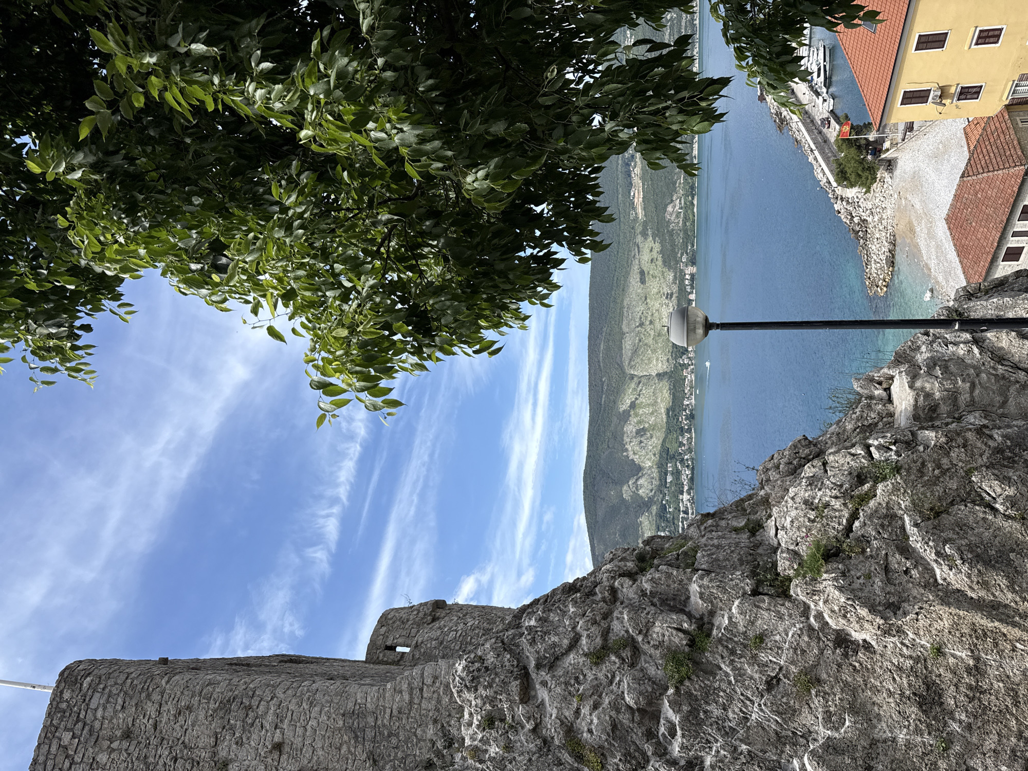 The image shows a view of Herceg Novi, Montenegro, from a high vantage point, likely the Kanli Kula fortress. The foreground features the textured stone wall of the fortress. Green foliage from a tree hangs overhead, framing the scene. In the distance, the blue waters of the Bay of Kotor stretch out, with a mountainous coastline visible across the bay. A lamppost stands near the fortress wall, pointing towards the water. Buildings with red-tiled roofs and a small harbor with boats are visible along the coastline. The sky is blue with wispy white clouds.
