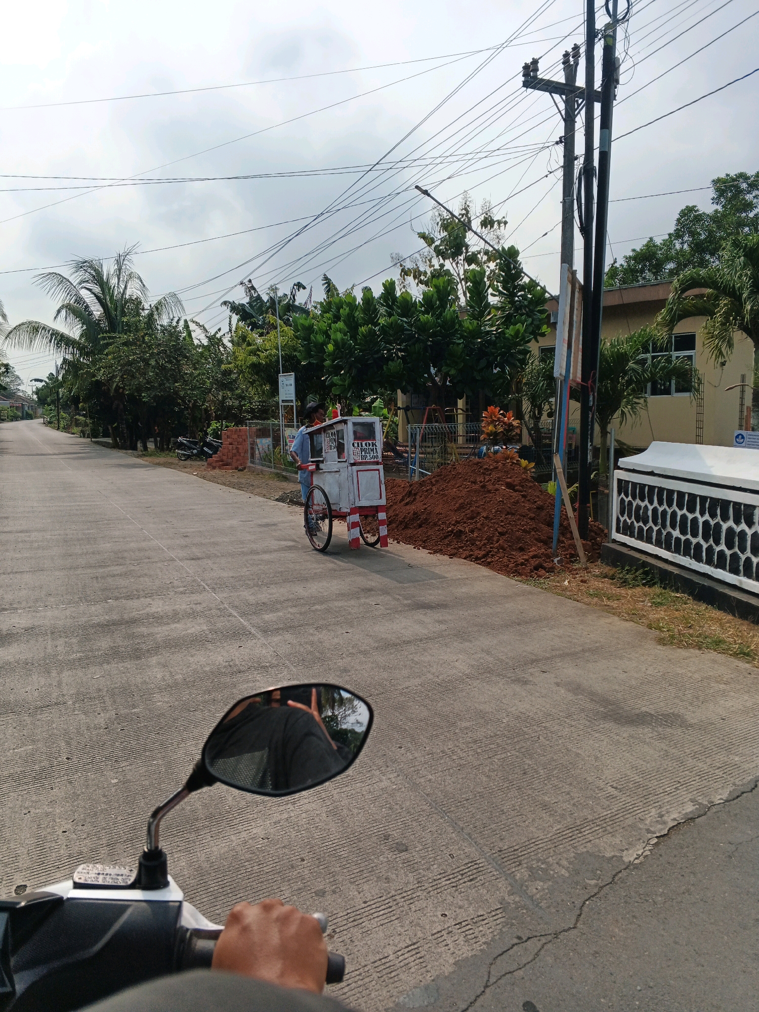 A person wearing a blue shirt and a dark cap is pushing a food cart on a paved road in Ciwuni, Indonesia. The food cart is white with red and white trim and has text written on it. The road is a two-lane asphalt road with textured concrete on the edges. There are power lines overhead, a telephone pole, and various trees and vegetation lining the road. A pile of dirt is visible on the right side of the road, next to a building with a white fence. In the foreground, a motorcycle handlebar and a mirror are visible, suggesting the photo was taken from a motorcycle. The sky is overcast with clouds.