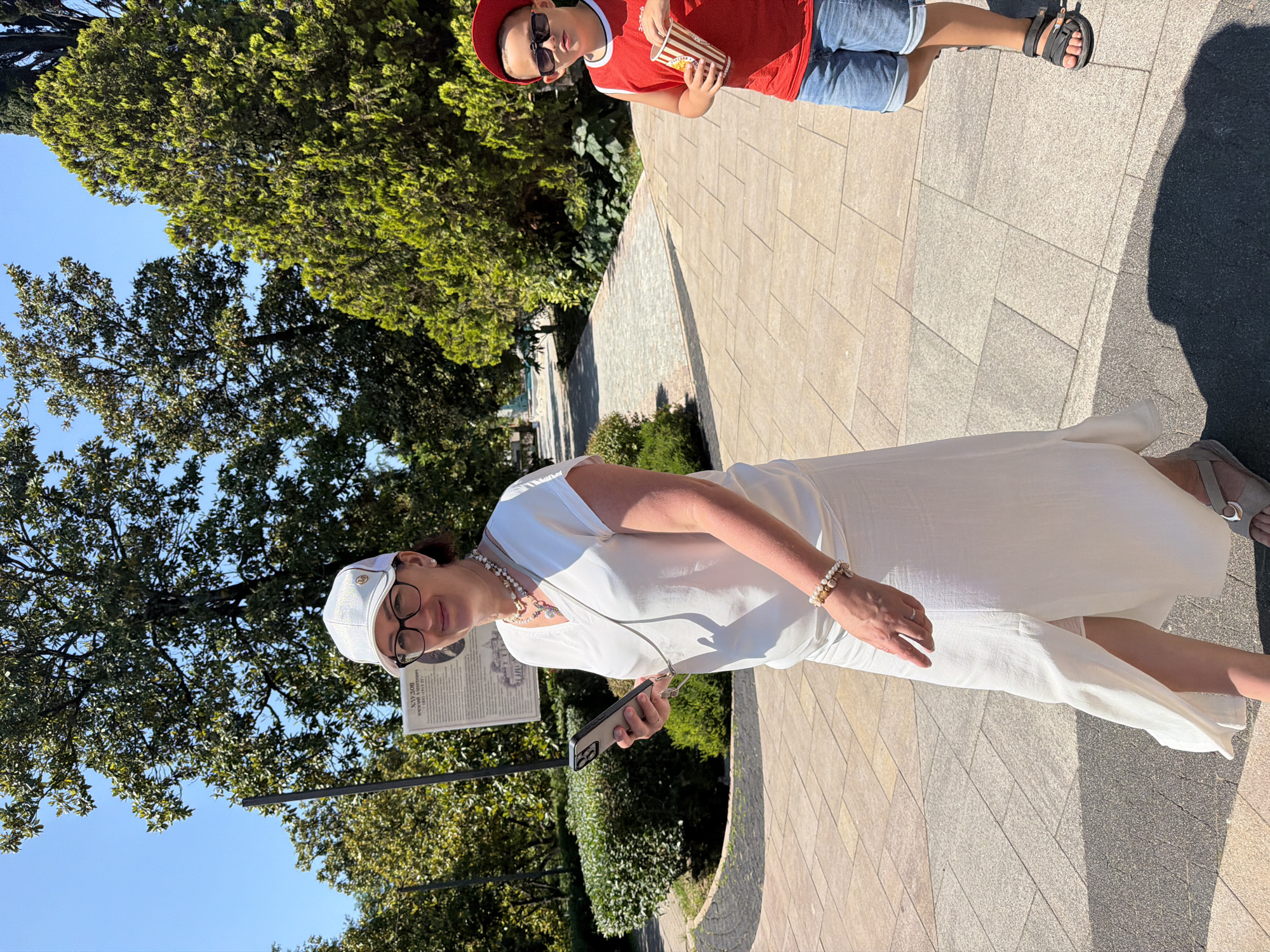 In the city of Sochi, Russia, on a sunny day, a woman and a young boy are walking on a paved pathway in a park. The woman, wearing a white cap, glasses, a white V-neck dress, and sandals, holds a smartphone in her left hand and is looking at it. She is adorned with several necklaces and a bracelet. The boy, to the right, wears a red sleeveless shirt, a red cap, sunglasses, denim shorts, and black sandals. He is holding a red and white striped cup of popcorn. Lush green trees and bushes surround the pathway. A informational signpost is visible behind the woman.