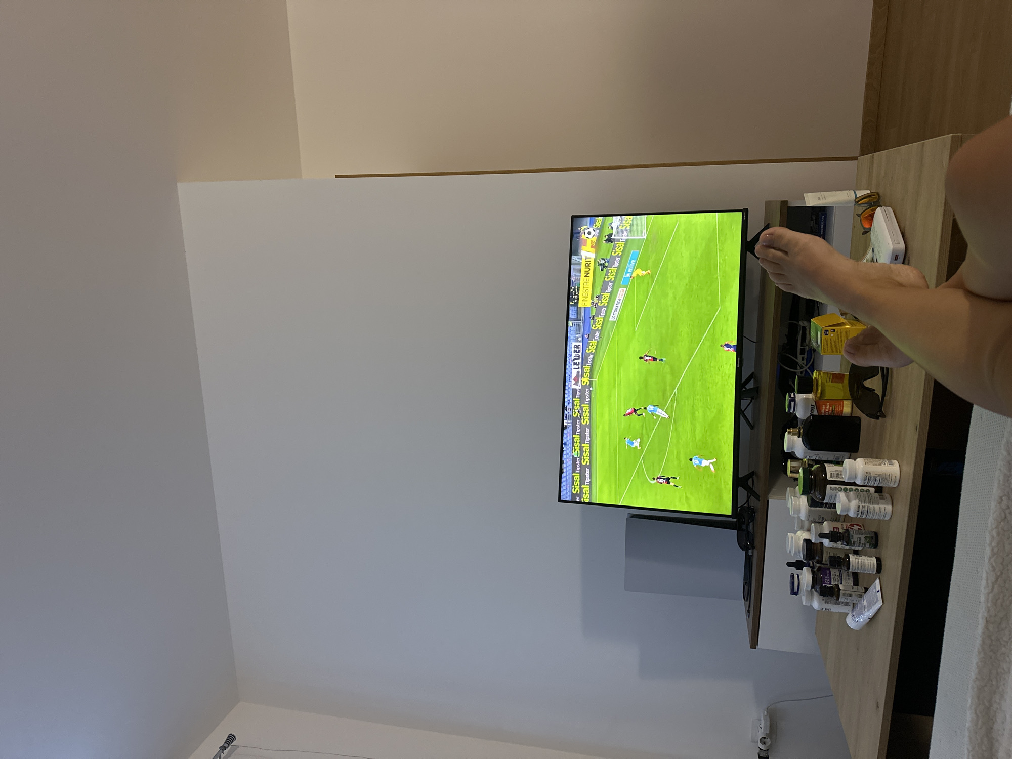 The image depicts a living room interior, likely in Donji Pobori, Montenegro, where a person is relaxing and watching a soccer match on a television. The television screen shows a live game with players on a green field and advertisements along the top border. In the foreground, a wooden table is covered with various bottles of supplements and medications, alongside a pair of sunglasses and other personal items. The person's legs and feet are visible, crossed and resting on the table, indicating a relaxed posture. The overall environment suggests a casual setting, possibly a home or an apartment.