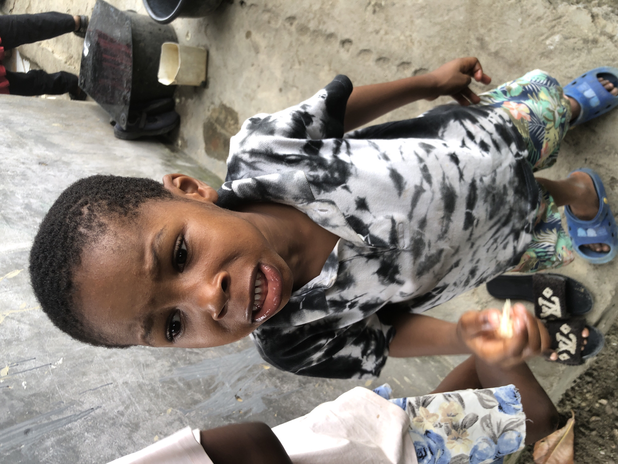 A young Black child, likely a boy, stands looking towards the camera with a distressed expression, mouth open showing teeth and gums. He is wearing a tie-dye t-shirt and floral shorts. His left hand is held out, holding a piece of light-colored food. He is barefoot, but wearing blue Crocs on his feet. Nearby, another child is partially visible on the left wearing a white shirt and floral shorts. The background appears to be an outdoor area with a concrete floor and a textured, grey wall. Two large black containers and a white plastic cup are visible in the background, along with the legs of an adult. The overall scene suggests a potentially impoverished setting in Ado, Nigeria.