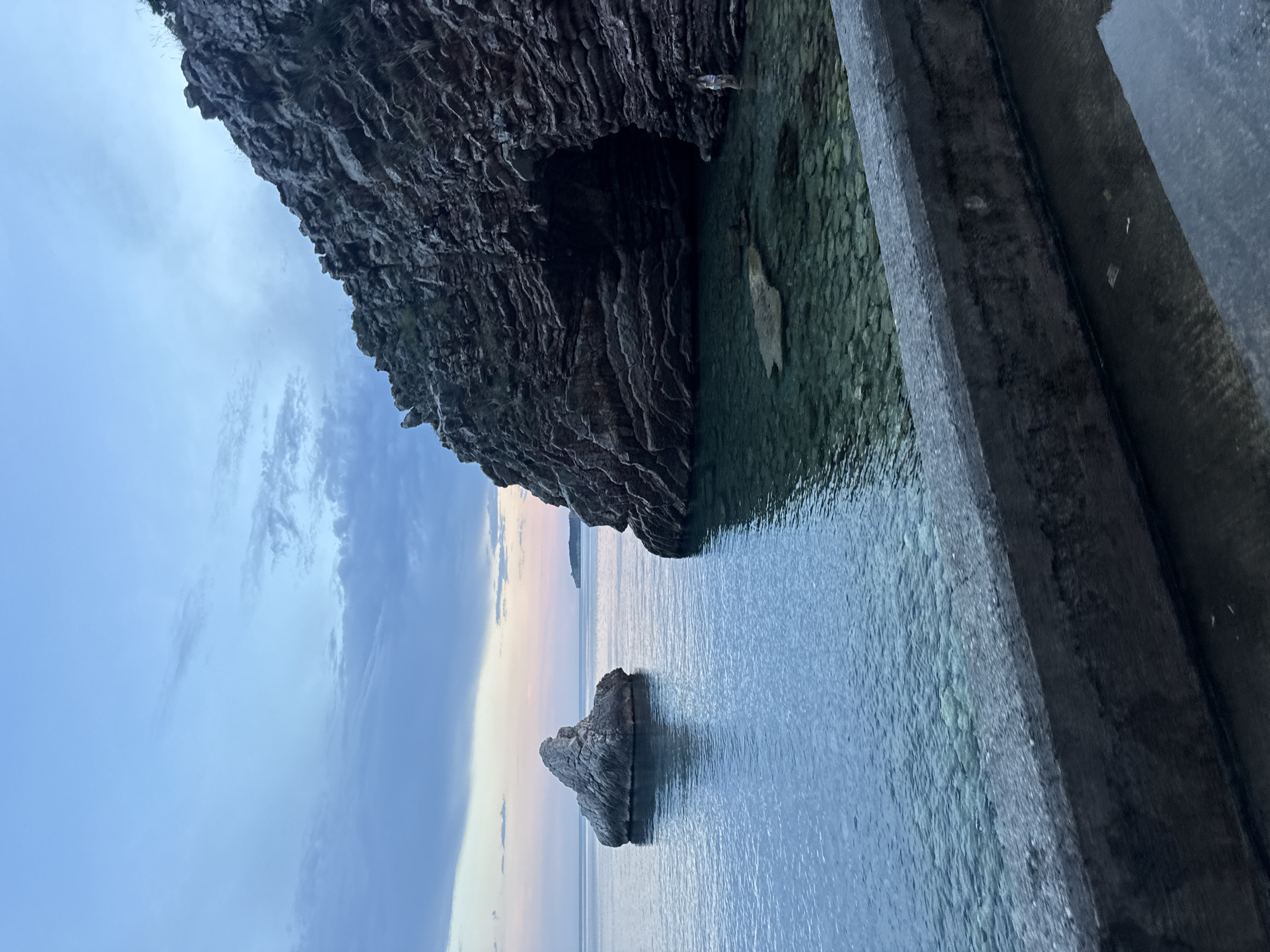 The media file captures a serene coastal scene at dusk in Rafailovići, Montenegro. The primary subjects are a large, rugged cliff face and the clear, calm water of the Adriatic Sea. The cliff, which appears to have a cave or overhang, dominates the upper left portion of the frame. A solitary, weathered rock formation juts out of the water in the foreground, adding a sense of scale and visual interest. To the right, a man-made concrete structure, possibly a retaining wall or part of a dock, runs along the edge of the water.
The time of day is clearly evident as twilight. The sky is painted with soft hues of blue, pink, and orange, indicative of the sunset or sunrise. Wispy clouds drift across the sky, adding texture and depth. The water is tranquil, reflecting the soft colors of the sky and the surrounding landscape. The overall mood is peaceful and contemplative, evoking a sense of natural beauty and stillness. No people are visible in the image, and there is no visible text. The location is specifically identified as Rafailovići, Montenegro. The scene suggests a quiet moment by the sea, ideal for reflection or appreciating the coastal environment.