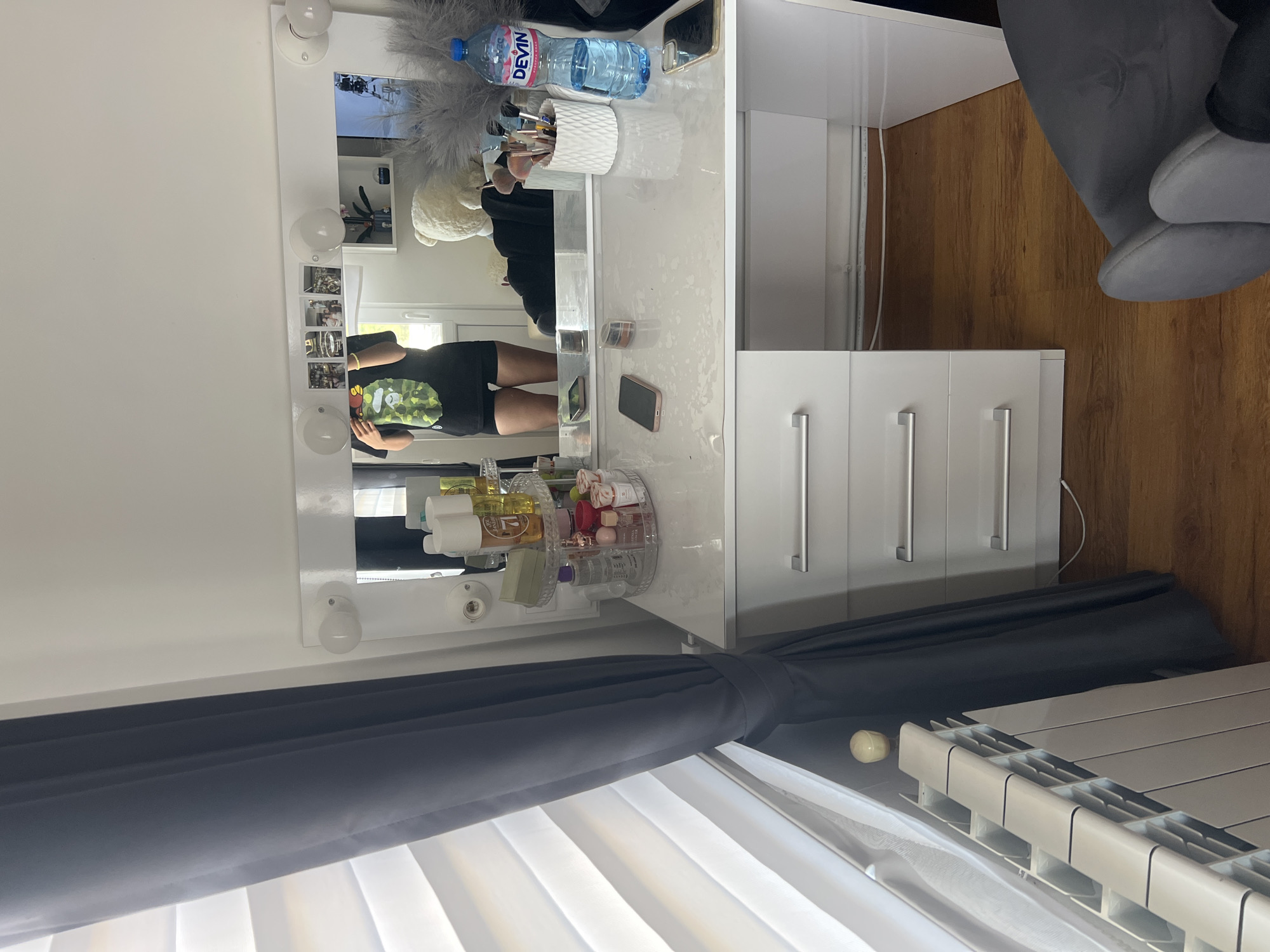 The image depicts a vanity table with a mirror, located in a room with white walls and a wooden floor. A person wearing a black shirt and shorts is reflected in the mirror. On the vanity table, there's a bottle of "Devin" water, makeup brushes in a white container, and various cosmetic products. The window is covered with a white blind and a gray curtain. A radiator is visible near the window. A gray chair is present in the room. The image does not provide enough information to determine the location in the city of Unknown, Unknown.