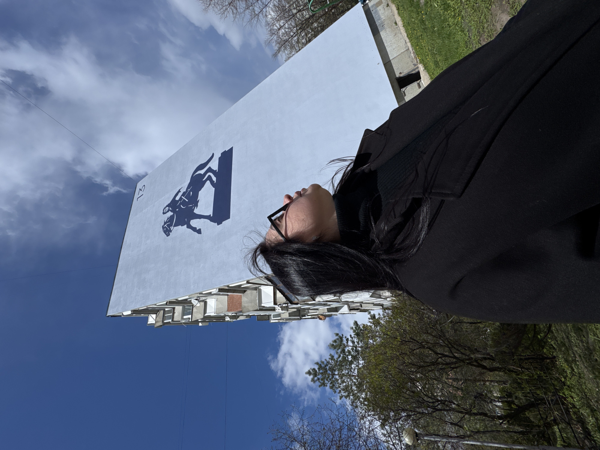 The image, originally rotated 90 degrees clockwise, depicts an outdoor scene under a bright sky. The primary subjects are a person and a large building with a mural.
A person with dark hair and glasses, wearing a black jacket, is visible in the foreground, positioned to the right side of the frame. The person is looking upwards, their face mostly in profile, seemingly observing the building or the sky.
The prominent object in the background is a tall, light-colored building, possibly an apartment block. A significant feature of this building is a large, dark blue mural painted on its side. The mural shows a dynamic silhouette of a figure on horseback, possibly a knight or warrior. To the left of this mural, the number "13" is clearly visible, also in dark blue. The building's side not covered by the mural reveals multiple windows.
The activity taking place appears to be the person observing their surroundings. The notable details include the time of day, which is daytime, indicated by the clear lighting and blue sky with scattered white clouds. The weather appears pleasant. In the background, patches of green grass and trees with some foliage are visible, suggesting a natural element in an urban or suburban setting. A thin wire or power line stretches across the sky in the upper left portion of the image. The visible text on the building is the number "13". The overall atmosphere is bright and open.