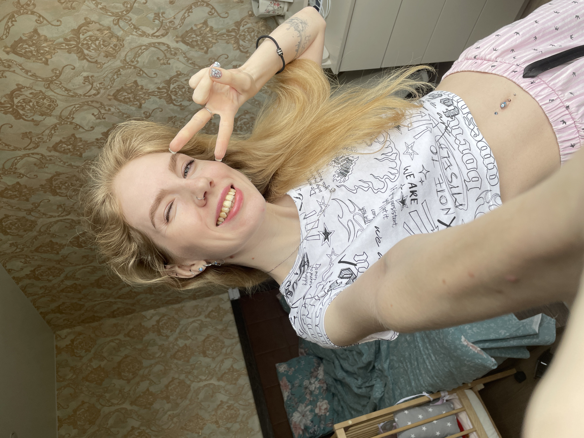 A young blonde woman is taking a selfie indoors, likely at home. She's lying down, turned slightly to the right, with her head at the top-left of the frame. She is smiling and winking with her right eye and holding up a peace sign near her face. She's wearing a white t-shirt with black drawings and text and pink and white striped pajama shorts. Her midriff is visible, showcasing a belly button piercing. She has several piercings in her ear and a nose ring.
The background features patterned wallpaper in shades of brown and tan. Part of a white piece of furniture is visible in the upper part of the frame. In the lower part, floral bedding and a baby crib are partly visible. A bracelet adorns her left wrist, and nail polish is visible on her fingernails.
There is no indication of a city or location, or any recognizable features to locate it in Izhevsk, Russia.