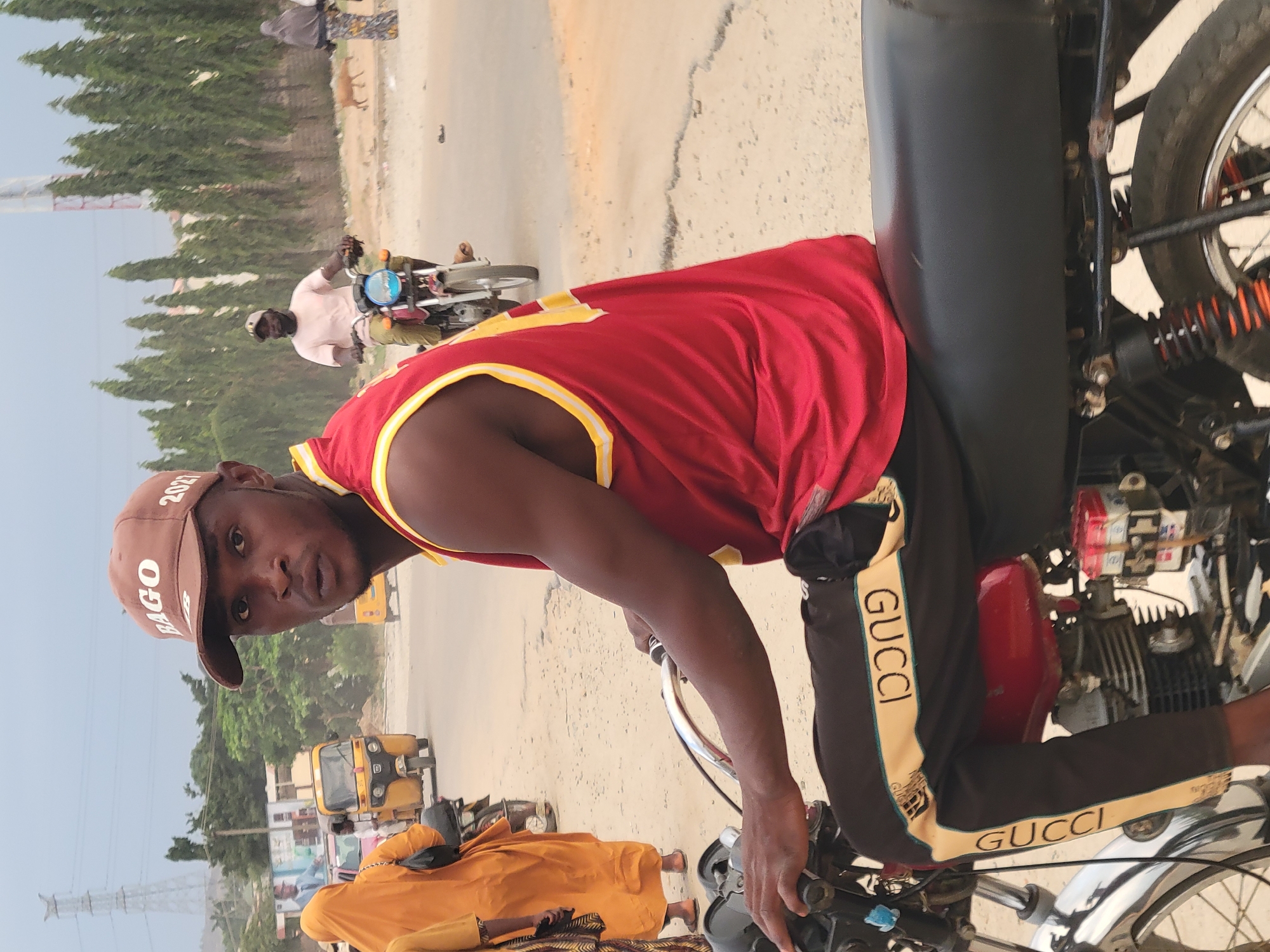 A man wearing a red tank top with yellow trim and dark pants with "GUCCI" printed in gold stripes is sitting on a motorcycle. He is also wearing a brown cap. The background shows an urban environment, including other motorists, three-wheeled vehicles, trees, and utility poles. The location is Madalla, Nigeria.