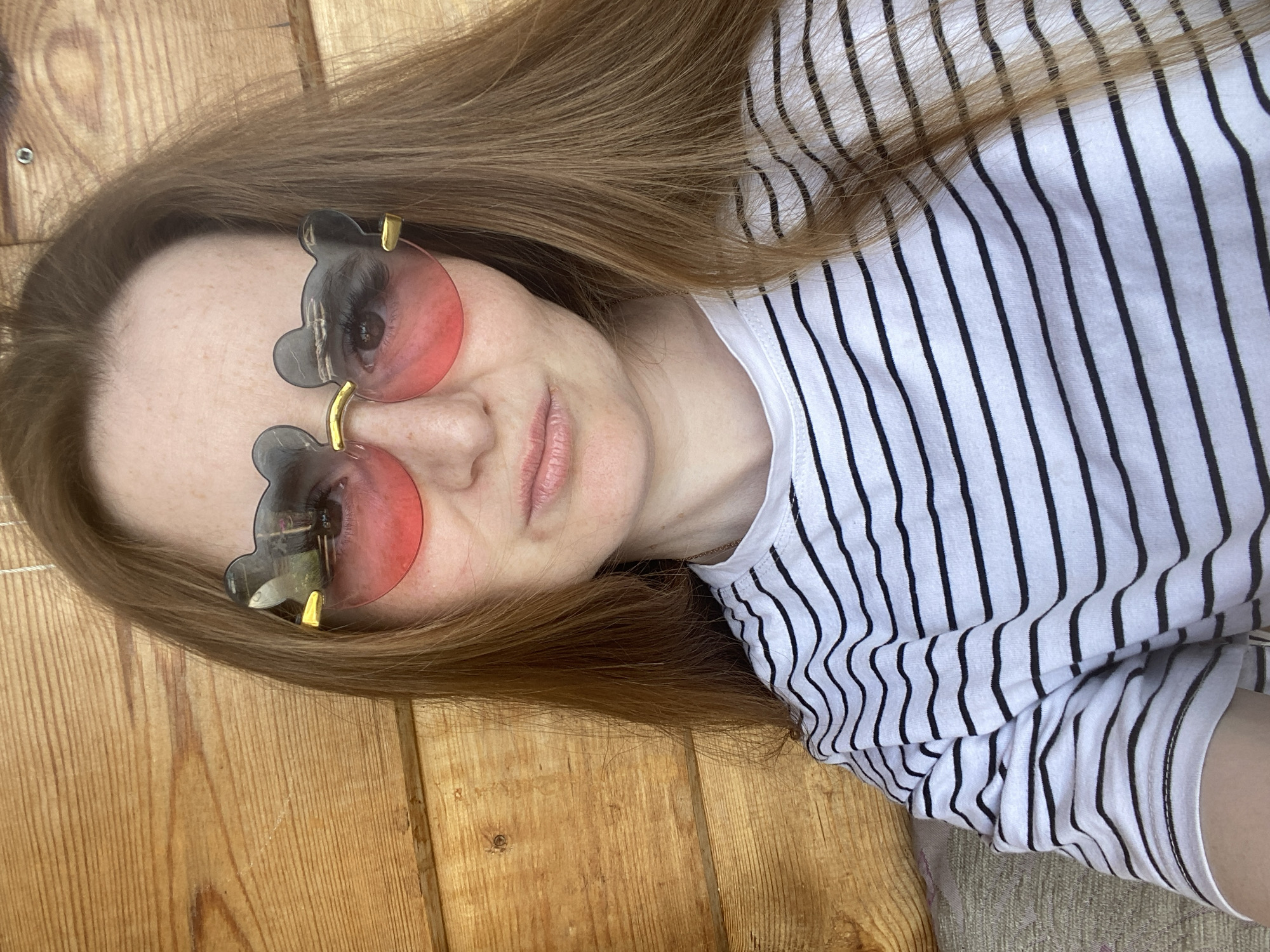 A person with long, light brown hair is lying down wearing bear-shaped sunglasses with pink lenses and a white T-shirt with black stripes. The background appears to be wooden planks. There is no information available to determine the location is the city of Chaikovsky, Russia.