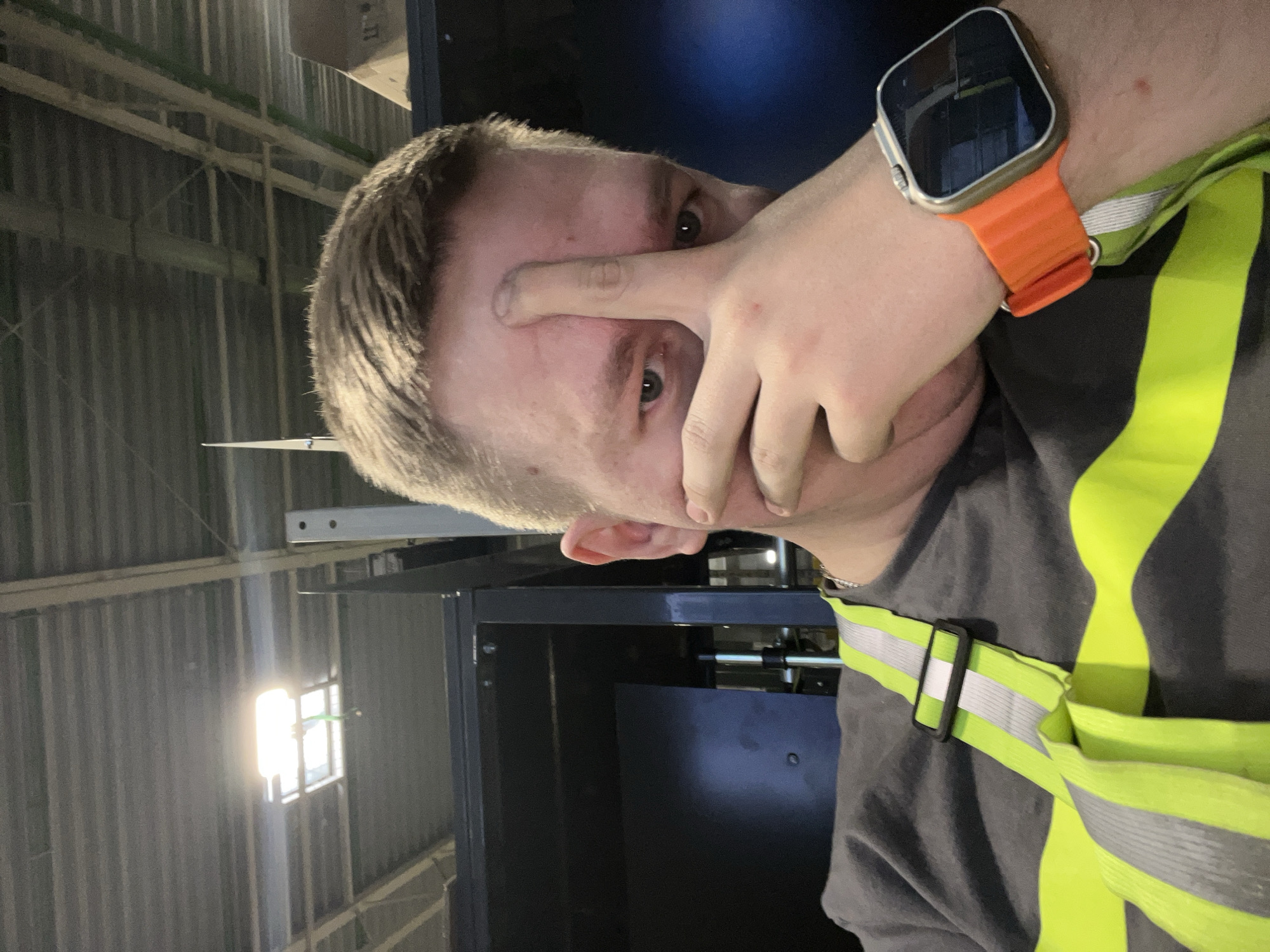 The image shows a close-up, selfie-style shot of a person's face and upper body. The individual has fair skin, short, light brown hair, and is looking directly at the camera. Their right hand is raised, with the thumb and index finger touching their forehead, obscuring part of their face. They are wearing a dark gray t-shirt with a bright yellow and silver reflective safety vest over it. On their left wrist, they are wearing a silver smartwatch with an orange band. The background appears to be an indoor industrial or warehouse setting with a corrugated metal ceiling and lighting fixtures. There are dark blue and black structures visible in the background. The image does not contain any information that would allow it to be definitively placed in the city of Lupanovo, Russia.
