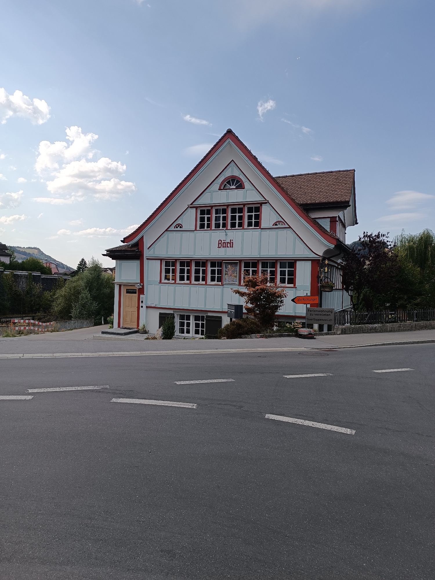The image depicts the exterior of a building identified as "Bärli" in Appenzell, Switzerland. The building is a multi-story structure with a distinctive red and light blue facade, characterized by numerous windows with red frames. A peaked roof with brown tiles crowns the building. The front of the building features a wooden door and a small garden area. To the right of the entrance, a signpost indicates directions to "alle Richtungen" (all directions) and a separate sign advertises "Ferienwohnung zu vermieten" (holiday apartment for rent) with the website "baerllappenzell.ch".
The building is situated alongside a paved road, indicated by white lane markings. Beyond the building and road, a fence encloses an area with various trees and foliage. In the background, rolling hills and mountains are visible under a partly cloudy sky. The scene appears to be a quiet street in a town or village, with no people or vehicles present. The overall impression is one of a well-maintained property in a scenic location.