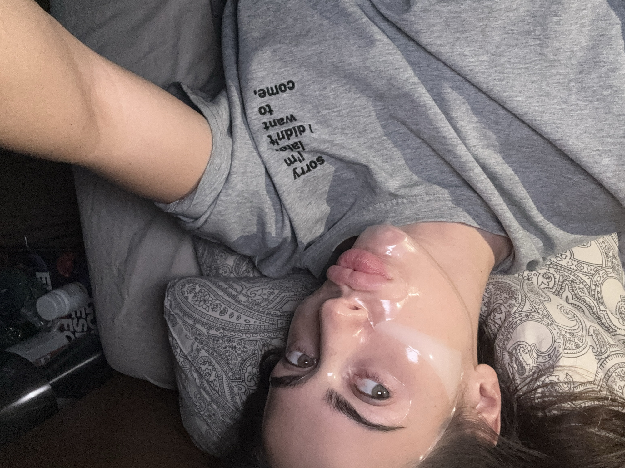A selfie shows a woman lying on a paisley pillow, wearing a gray t-shirt with the words "sorry i'm late, i didn't want to come" printed on the sleeve. A clear, glossy substance covers her face. Other items are visible in the lower left corner of the frame. There is no indication of the location in Warsaw, Poland.