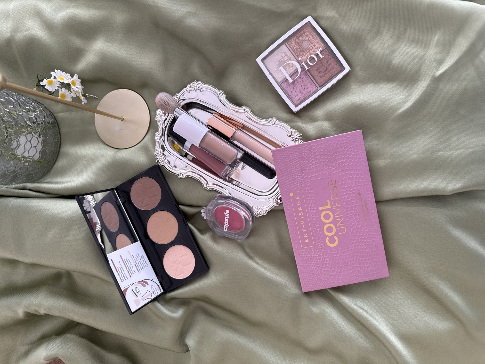A collection of makeup products is arranged on a wrinkled, pale green satin cloth. In the upper left corner, an open makeup palette with three circular pans of powder is visible, with instructions printed inside the lid. To the right of the palette is a small, clear plastic capsule containing a pink powder. Below this is a rectangular pink eyeshadow palette with gold lettering that reads "ART-VISAGE COOL UNIVERSE." To the right of the pink eyeshadow palette and slightly above it, a small silver tray holds several lip glosses and a makeup brush. In the bottom right corner, a square Dior eyeshadow palette with four circular pans of eyeshadow is placed on the cloth. A decorative glass vase with a metallic honeycomb pattern is visible in the upper right background, next to a small white daisy floral arrangement on a gold stand. The lighting is soft and even, suggesting an indoor setting, possibly a vanity or dressing table. There is no indication of the location being Izhevsk, Russia, nor any people, actions, or interactions.