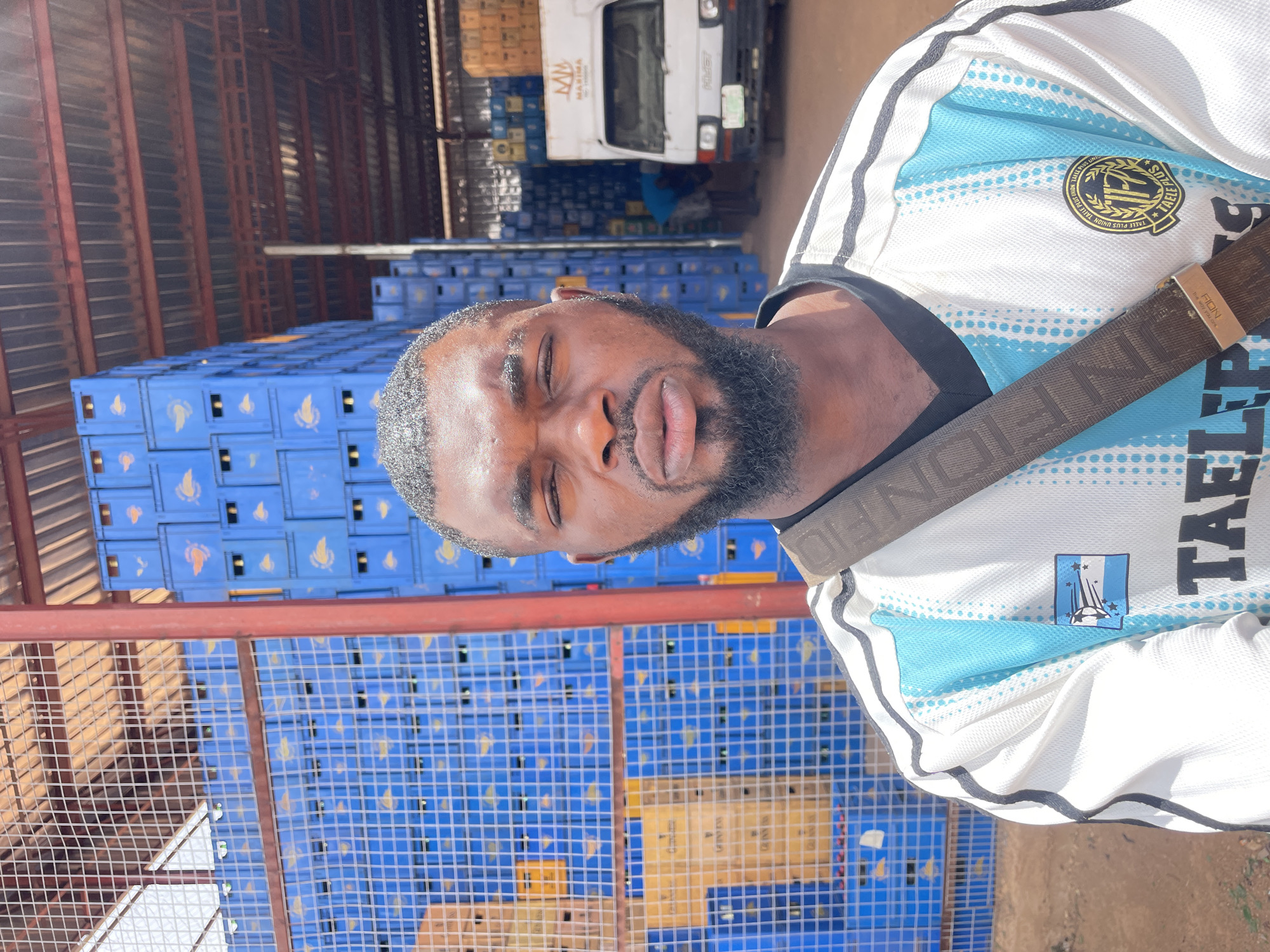 A close-up, slightly askew, daytime shot features a man with dark skin, a full beard, and a short haircut, positioned in front of stacked blue crates. The man is wearing a white t-shirt with light blue accents and a dark strap crossing his torso. Text and logos are visible on the shirt.
Behind the man, there are stacks of blue crates, suggesting a storage or industrial setting. Red metal support structures are visible between the crates. In the background, a white truck is partially visible.
Based on the OCR, the location is Gura Top, Nigeria.
