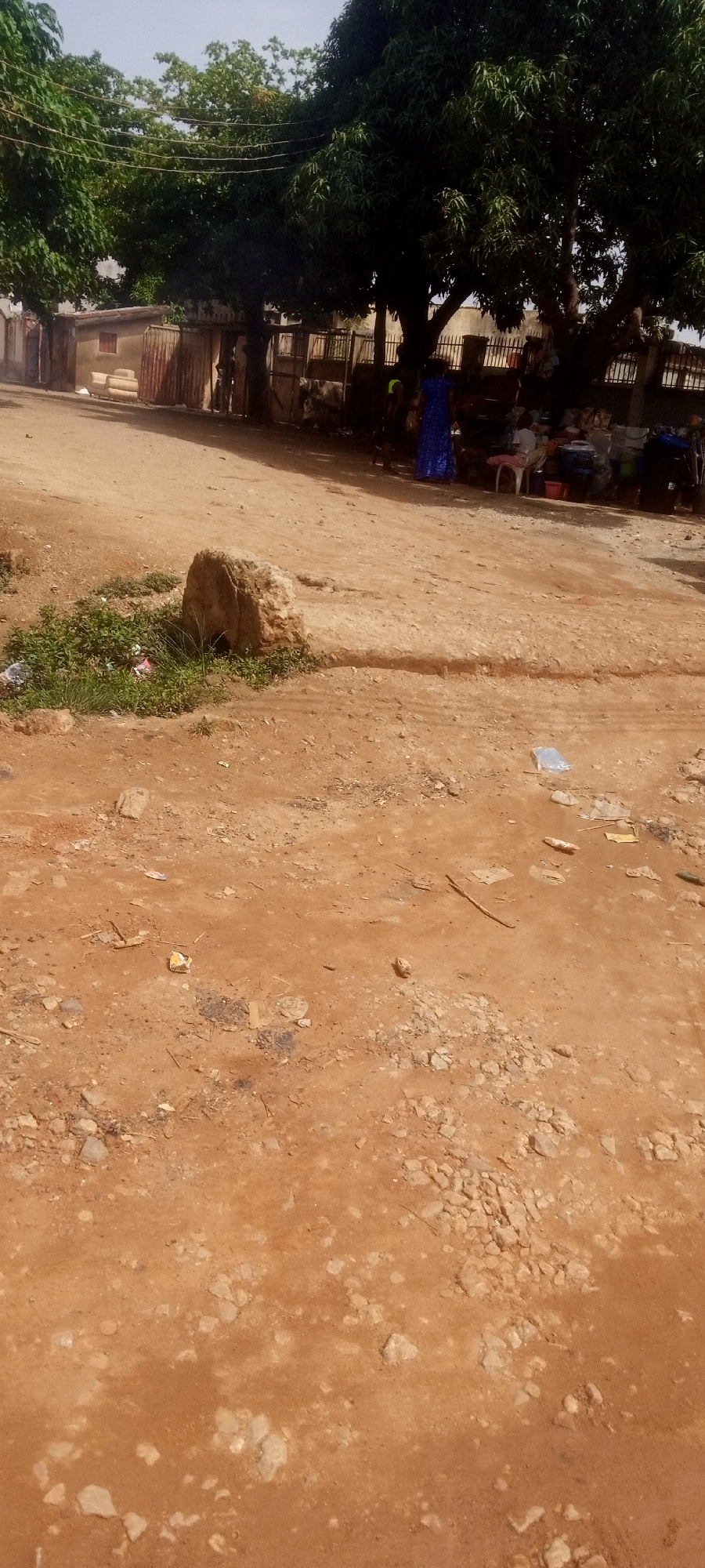 Yelwa, Nigeria: An unpaved, reddish-brown dirt road runs through a village. A large rock sits on a small embankment, with weeds growing nearby. Litter, including plastic bags, is scattered on the ground. Several people are gathered in the background under a row of trees, near a fenced building. One individual is wearing a bright blue dress. Objects, possibly for sale, are visible on tables. Overhead wires are present.