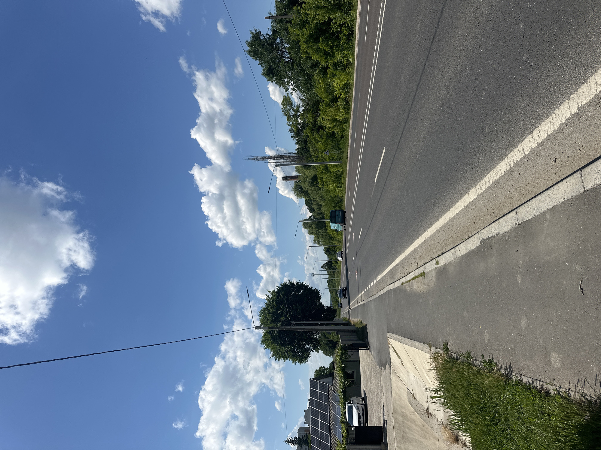 The image depicts a street scene in Strășeni, Moldova on a sunny day with a bright blue sky and scattered white clouds. The foreground shows a paved sidewalk with a grassy verge and a concrete drainage channel on the left. A single dark power line stretches diagonally across the frame from the top left.
The main focus of the scene is a two-lane asphalt road with white lane markings. Several vehicles are visible on the road: a light-colored car, a dark-colored car, a teal van, and two other cars further in the distance.
On the left side of the road, there is a house with solar panels on its roof, a fence, and a large, leafy tree. Beyond the house, more trees are visible. On the right side of the road, a dense line of trees and vegetation forms a natural backdrop. In the distance, industrial structures, including a chimney-like structure and tall poles, are present within the treeline. The overall environment suggests a semi-urban or suburban setting.