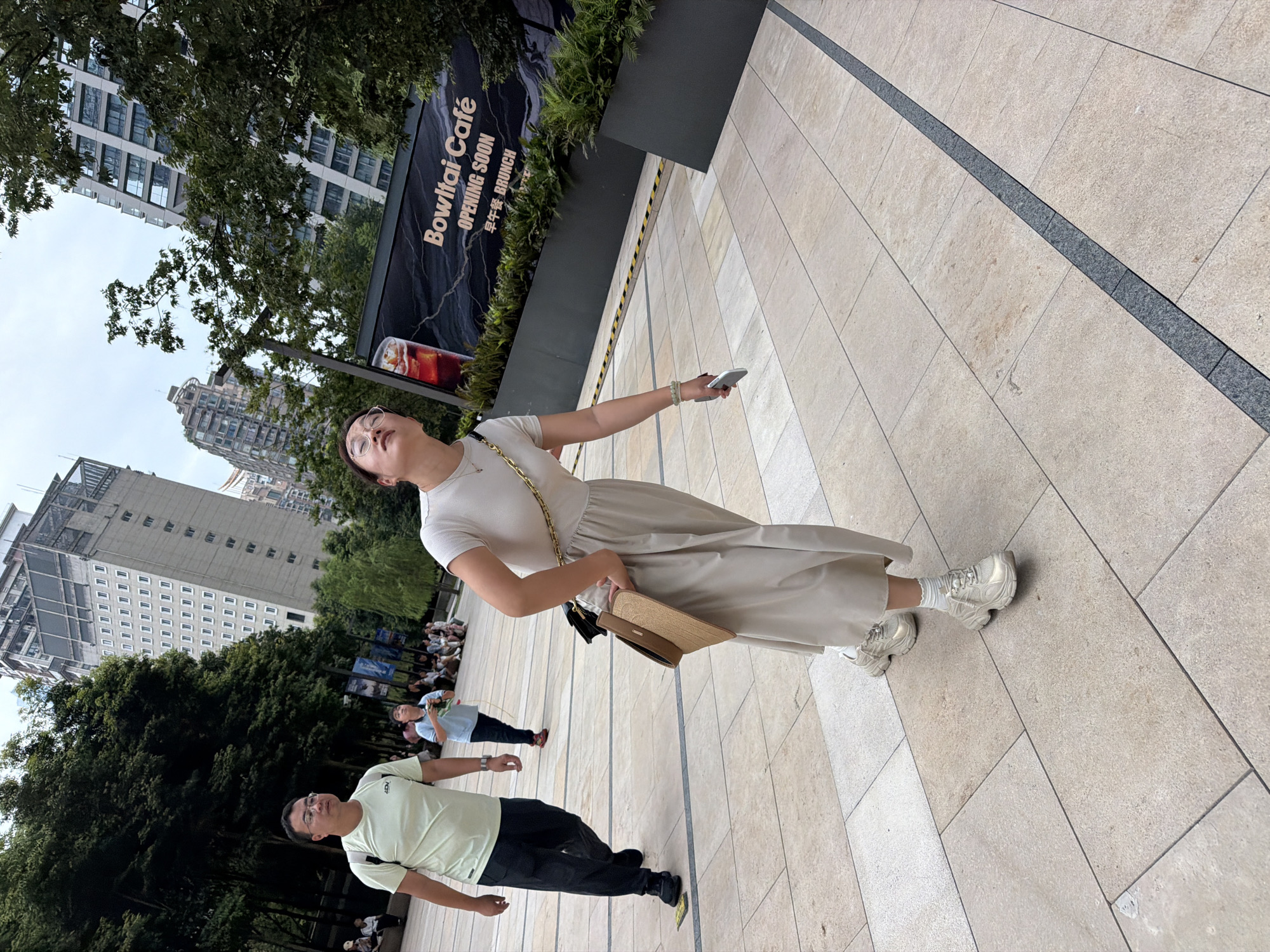 In the Yuzhong District of China, a street scene unfolds. A woman, wearing glasses and a light-colored dress, is captured in mid-stride, looking upwards with her mouth slightly open. She carries a woven purse on her left shoulder and holds a smartphone in her right hand. Her attire includes white socks and chunky, light-colored sneakers. To her left, a man in a light green t-shirt and dark pants walks in the opposite direction, his gaze directed forward. He wears glasses and carries a backpack. In the background, several people are seated, and a young child in a blue shirt is visible. A large banner advertising "Bowital Café" with "OPENING SOON" and "BRUNCH" in English and Chinese is displayed. The environment features a paved walkway with tiled sections and a dark gray stripe running through it, bordered by lush green foliage and modern, multi-story buildings. The sky is overcast.
