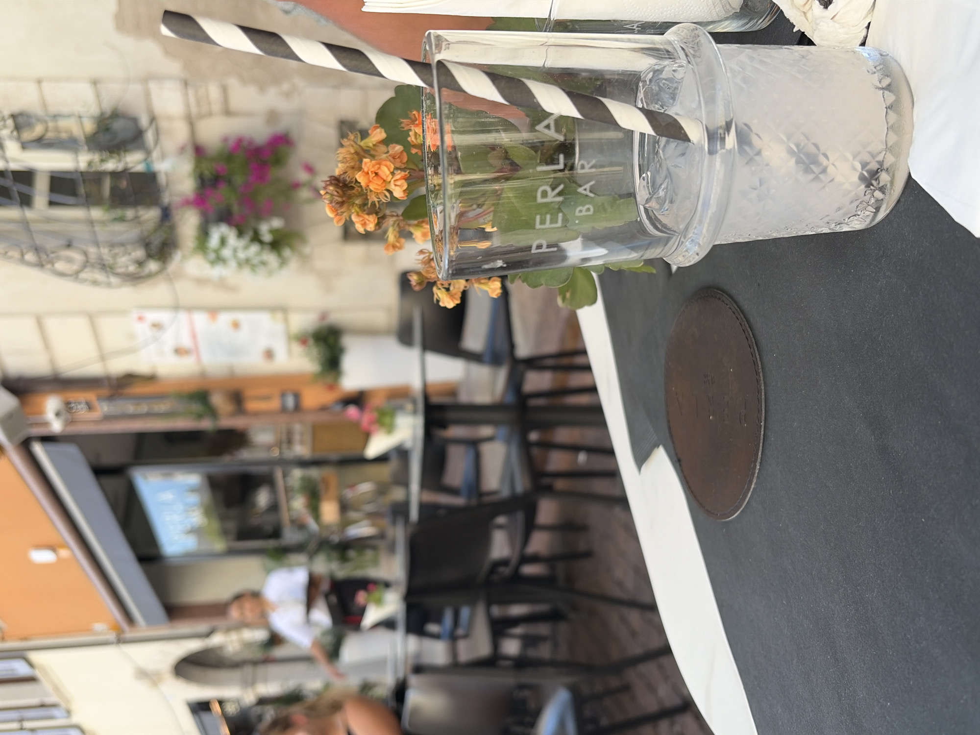 The image depicts an outdoor cafe scene in Trento, Italy, with a focus on a table in the foreground. A clear glass filled with a light-colored beverage and ice sits on the table, adorned with a black and white striped straw and a small orange flower arrangement. The glass has the branding "PERLA BAR" etched onto it. A dark circular coaster is visible to the left of the glass. In the background, out of focus, are tables and chairs, patrons, and the facade of a building. The overall setting suggests a warm, sunny day.