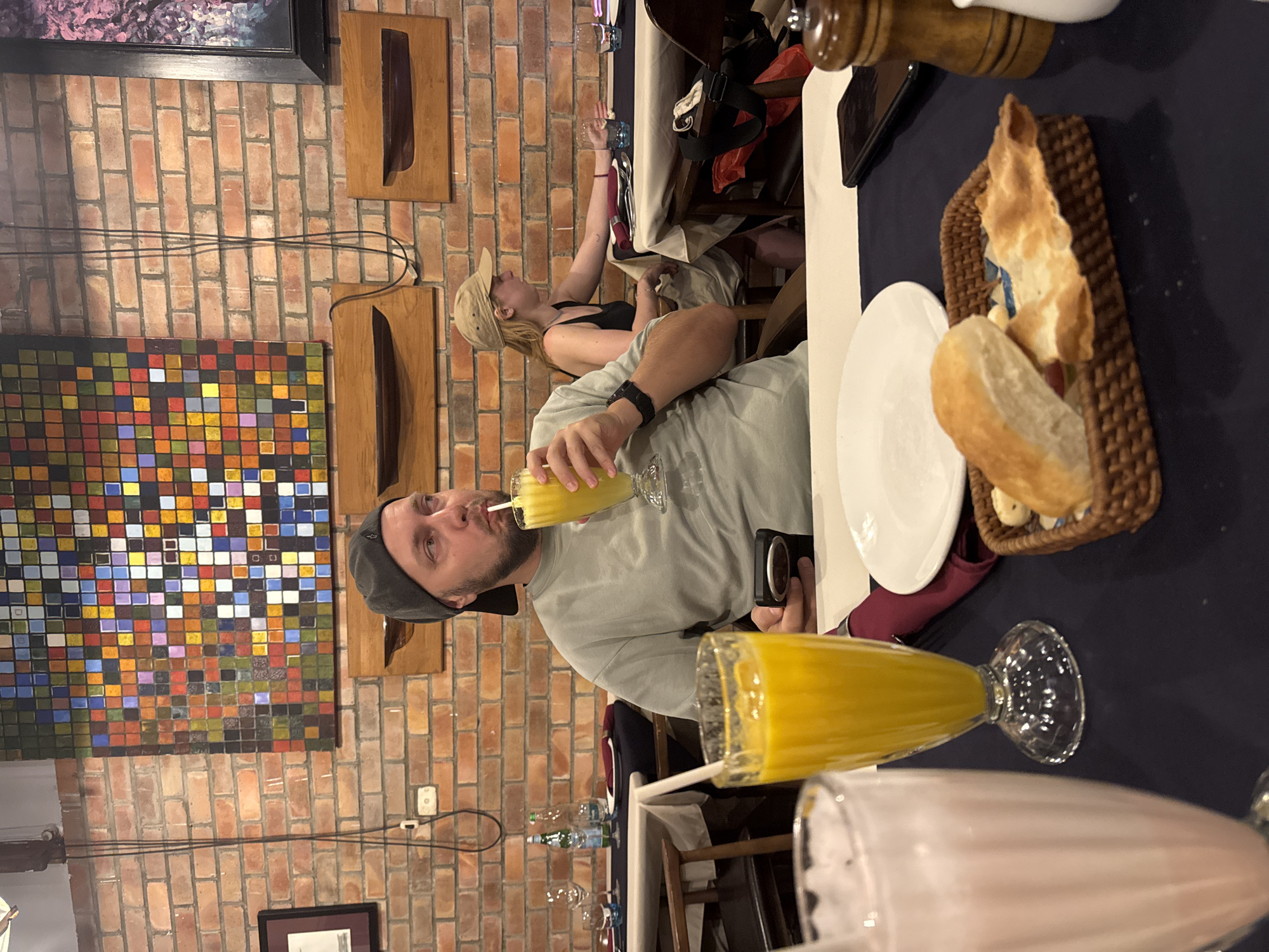 A group of people are sitting at a table in a restaurant in Hà Nội, Vietnam. A man is drinking a yellow smoothie through a straw. On the table, there is a plate, a basket of bread, several drinks and glasses. The restaurant has a brick wall with colorful artwork and wooden decorations.