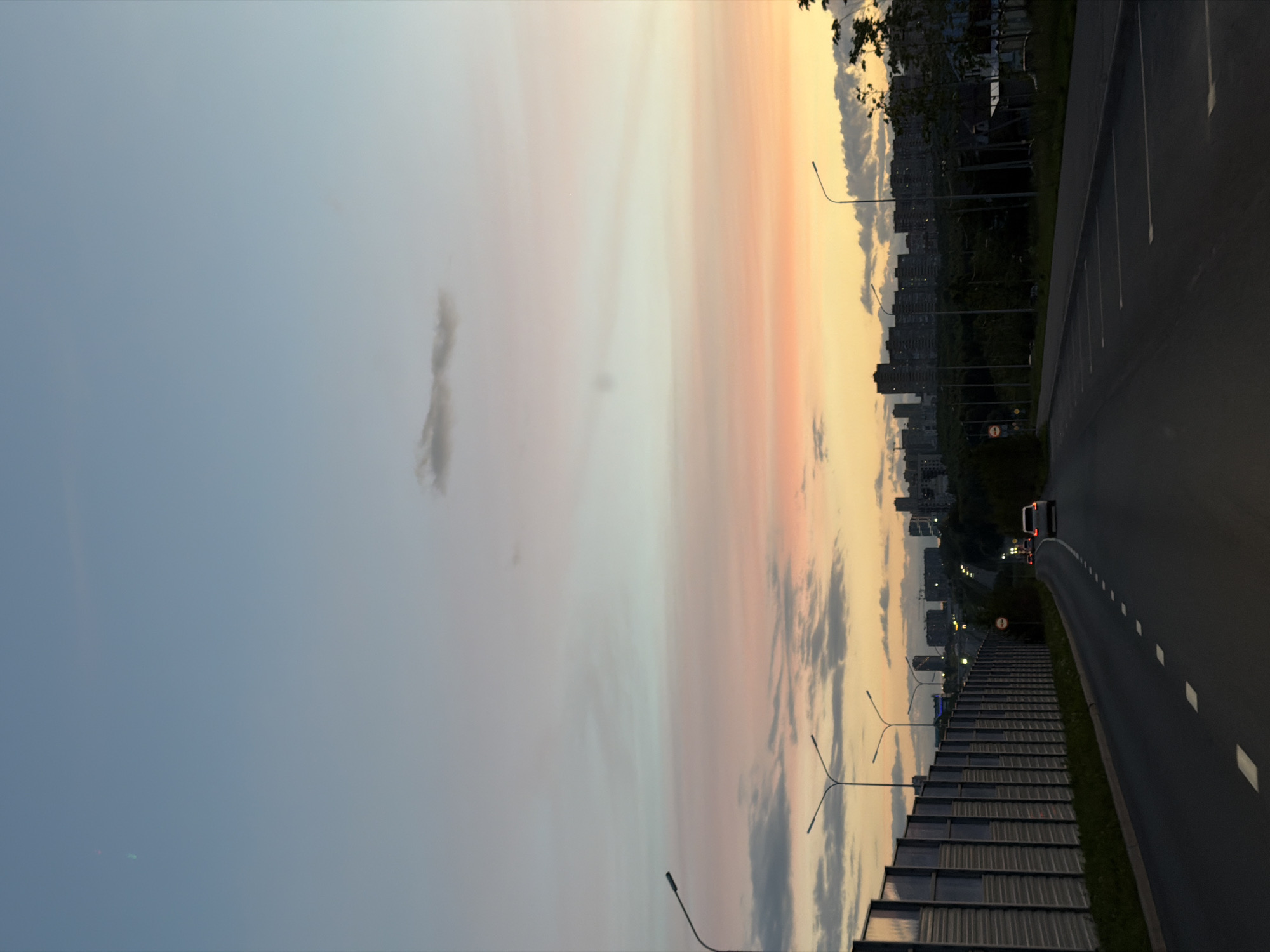 This media file captures a road leading towards a city skyline during a sunset. The scene is oriented vertically, with the sky occupying the left half of the frame and the road and city on the right. The sky displays a gradient of colors, transitioning from a pale blue at the top to soft pinks and oranges near the horizon, indicating it is either sunrise or sunset. Scattered clouds add texture to the sky.
Along the right side of the frame, a paved road curves away from the viewer, marked with white dashed lines. To the left of the road, there appears to be a tiered structure with glass panels, possibly a pedestrian walkway or a modern architectural element. A few streetlights are visible along this structure.
In the distance, a city skyline is silhouetted against the colorful sky. Tall buildings suggest an urban environment. A white van is seen driving on the road ahead, with its taillights illuminated, suggesting it is moving away from the viewpoint. The overall impression is one of a peaceful urban evening.
The time of day is clearly sunset due to the warm colors in the sky. The weather appears clear apart from the clouds. There is no visible text in the image. The location context provided is Kazan, Russia.