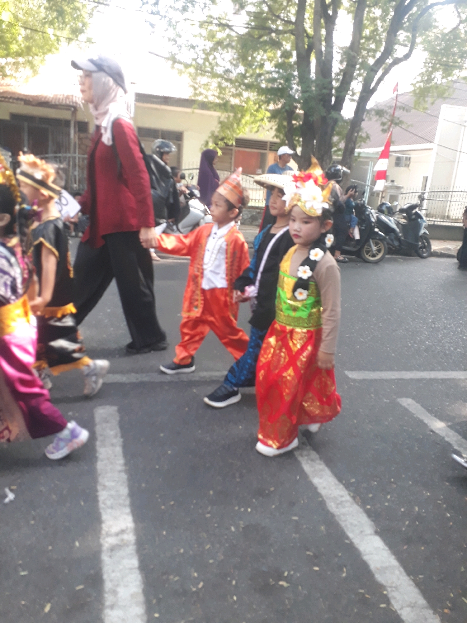In Pangkalpinang, Indonesia, a group of children dressed in traditional attire are participating in a parade. The children are walking on a paved road, with trees lining the background and buildings visible. Several adults are accompanying them, observing the parade. Motorcycles are parked on the side of the road. The scene suggests a cultural event or celebration.