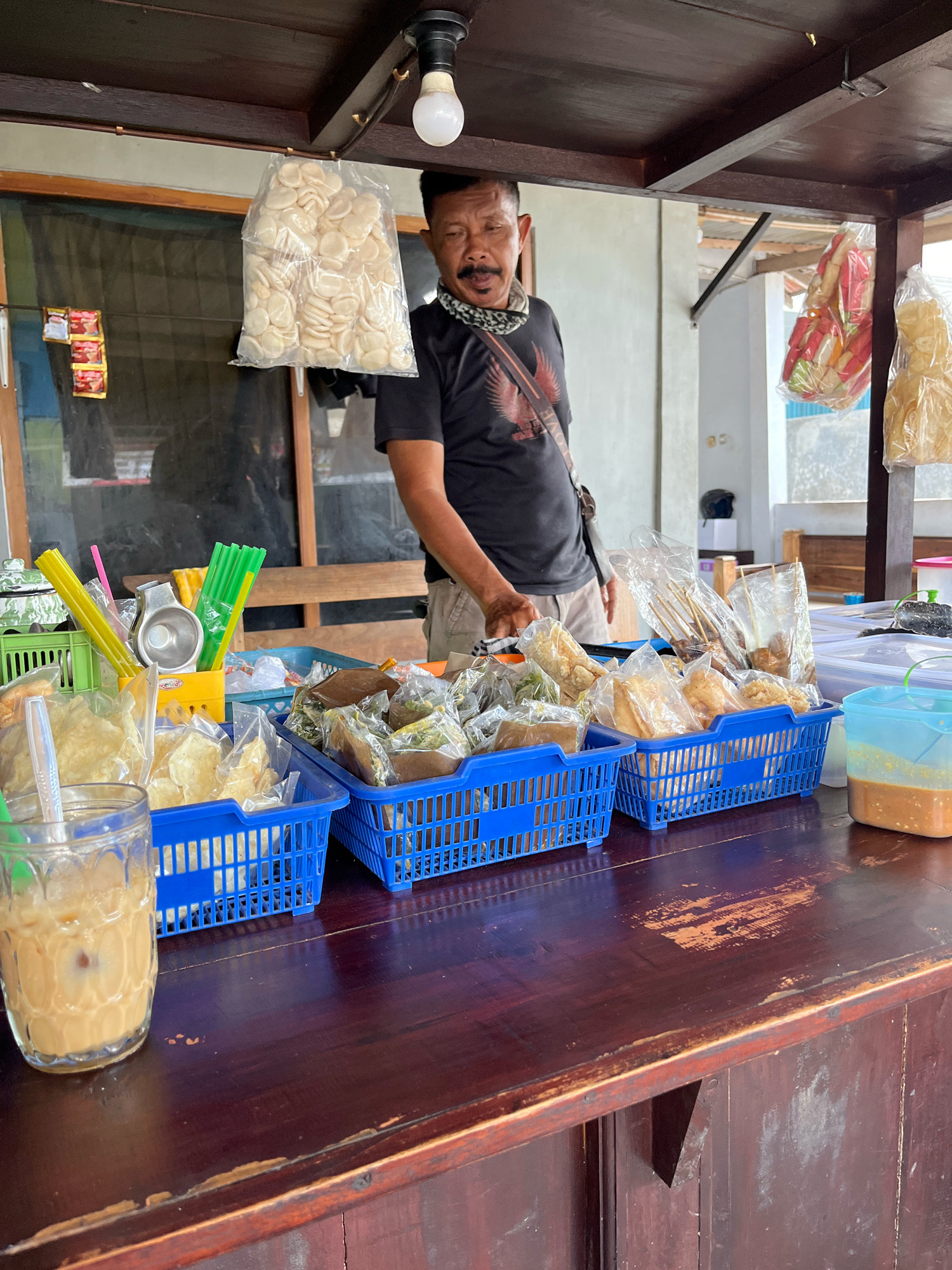 A middle-aged man with a mustache and a black t-shirt stands behind a wooden counter in Banguntapan, Indonesia, at a food stall. He wears a patterned bandana around his neck and has a sling bag across his chest. The stall is stocked with various food items packaged in plastic bags and displayed in blue plastic baskets. Among the items are what appear to be fried crackers, skewers of food, and other snacks. To the left of the counter, a glass filled with a light-colored beverage, likely iced coffee or tea, sits next to a green plastic basket containing yellow and green straws and a metal cup. Behind the man, bags of white, circular crackers hang from the ceiling. The stall appears to be outdoors, with natural light illuminating the scene. The wooden counter shows signs of wear and tear.