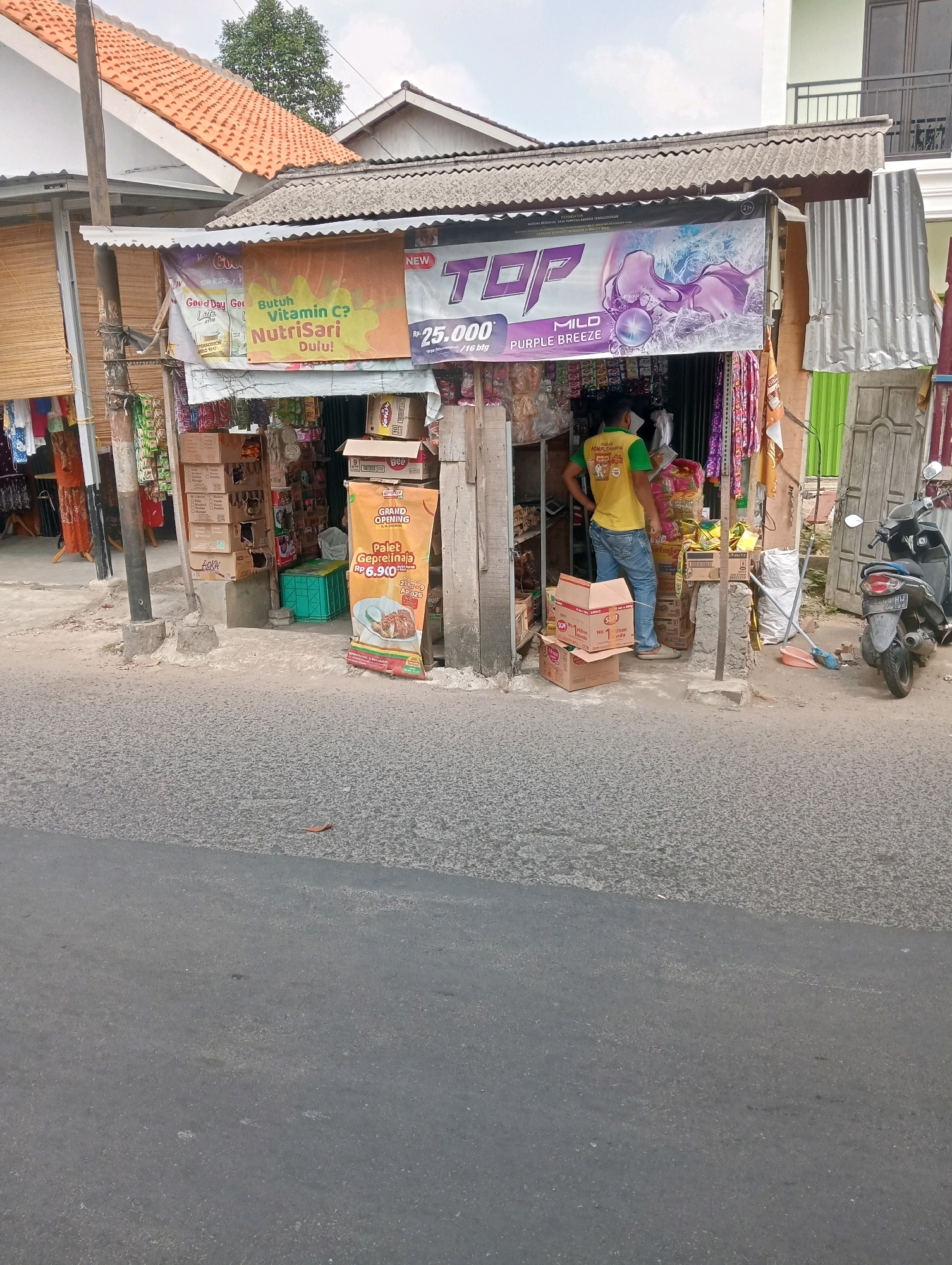 The image shows a street-level view of a small shop in what appears to be an urban or semi-urban environment. The shop is modest, with a corrugated metal roof and a facade made of wood and other building materials. In front of the shop, a variety of goods are displayed, including boxes of what appear to be packaged food or drinks. A large orange banner on the right side of the shop advertises a "Grand Opening" and offers a special deal on "Palet Geprejna" for RP 6.900. A man wearing a yellow t-shirt and blue jeans is standing inside the shop, appearing to be interacting with the merchandise or a customer. A black motorcycle is parked on the right side of the image, near the shop's entrance. The street in front of the shop is paved and shows signs of wear. The overall impression is of a local, informal retail establishment. The environment is outdoors, with a partially visible building in the background and a clear sky overhead.