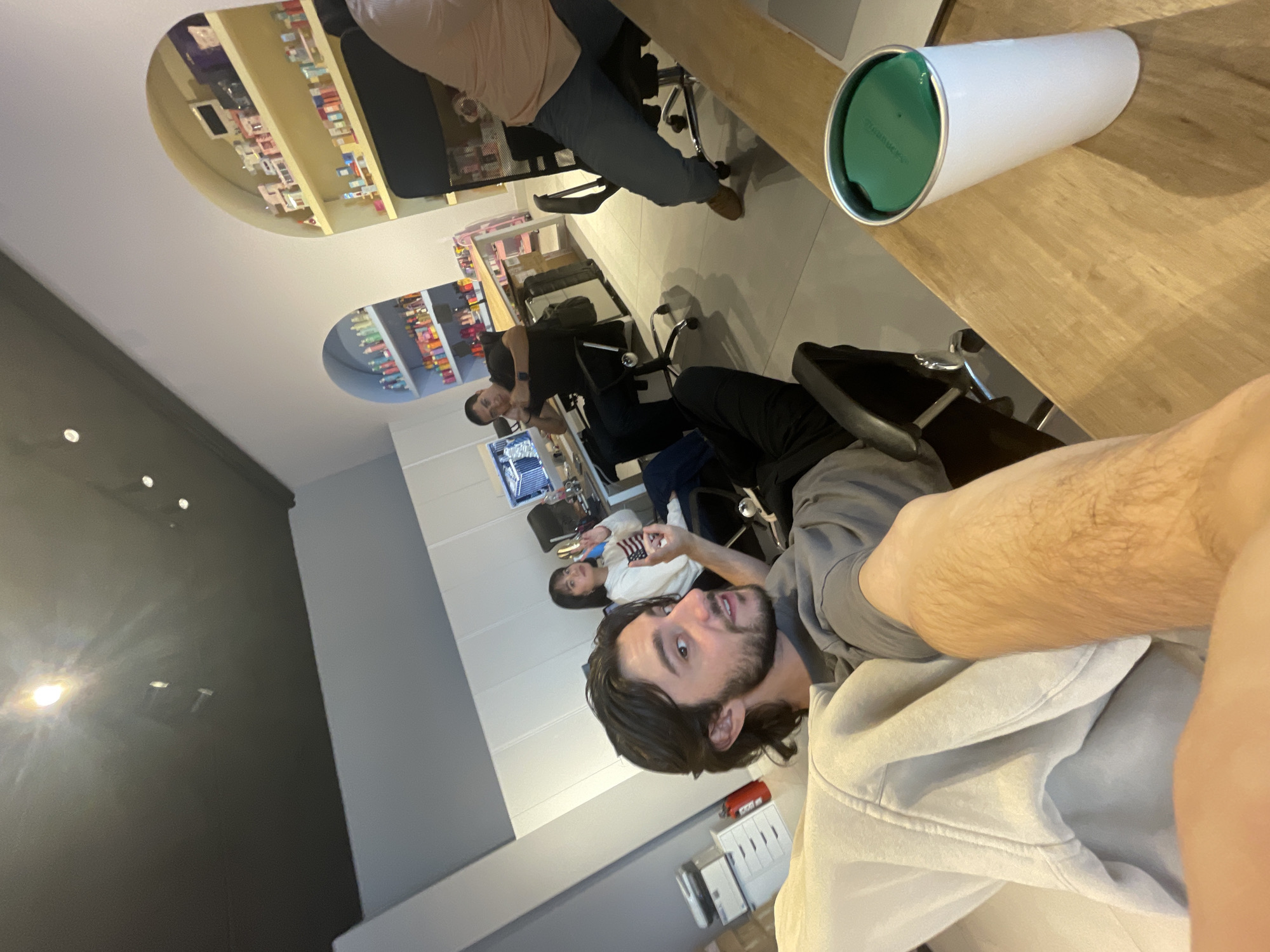 The image appears to be a selfie taken from a high angle inside a retail location with other people present. The location may be a pharmacy or health and beauty store based on the shelving filled with products visible in the background, which shows two reflections, indicating a mirrored surface. Several people are present and seated on rolling chairs at a long, presumably wooden, table. The foreground shows a person in a light-colored hoodie. A white and green to-go cup sits on the right of the table, and a computer screen is visible among the seated customers. While it's impossible to confirm the location without additional information, the store layout suggests a retail setting in Mexico City.