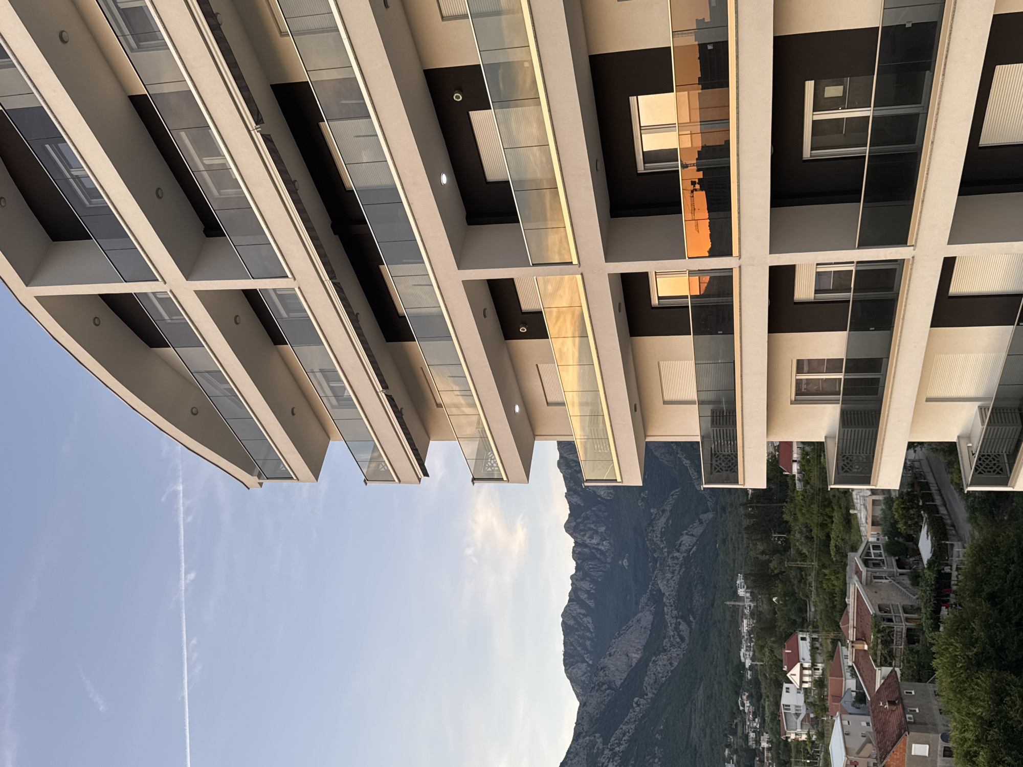The image depicts a modern apartment building in Šušanj, Montenegro, with a backdrop of lush green mountains and a clear sky. The building features multiple balconies with glass railings, reflecting the warm hues of the setting sun. Below the apartment building, a village with red-tiled roofs and scattered trees is visible. The foreground is dominated by the building's facade, showcasing its contemporary architecture. No people are visible in the scene. The overall atmosphere is tranquil, with the natural landscape contrasting with the urban development.