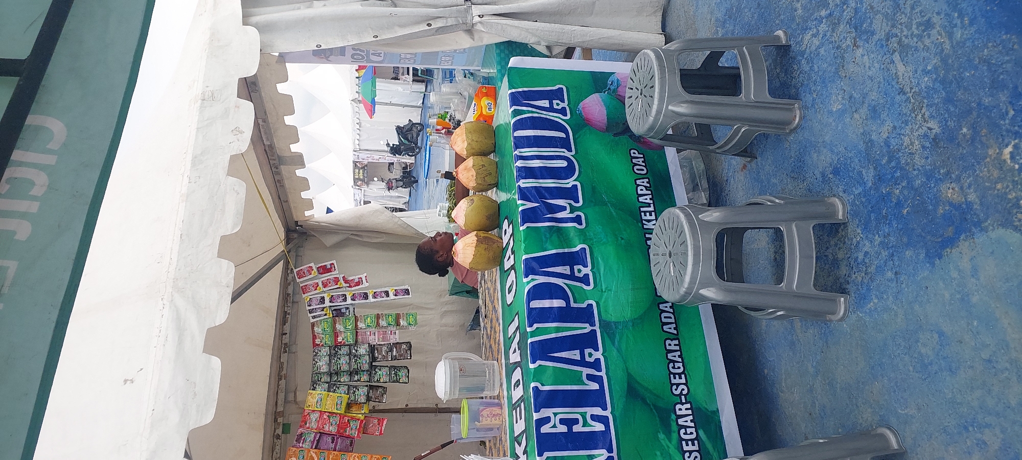 The image shows a street-side stall selling young coconuts in Asei Besar, Indonesia. A woman with dark hair tied back sits behind a table displaying four young coconuts and various packets of snacks or drink mixes hanging on the wall behind her. She is wearing a pink top. In front of the table, two gray plastic stools are placed on a blue-painted ground. A green banner with white and blue text, including "KELAPA MUDA," is stretched across the front of the table. The stall is covered by a white tent. In the background, other tents and a motorcycle are visible.