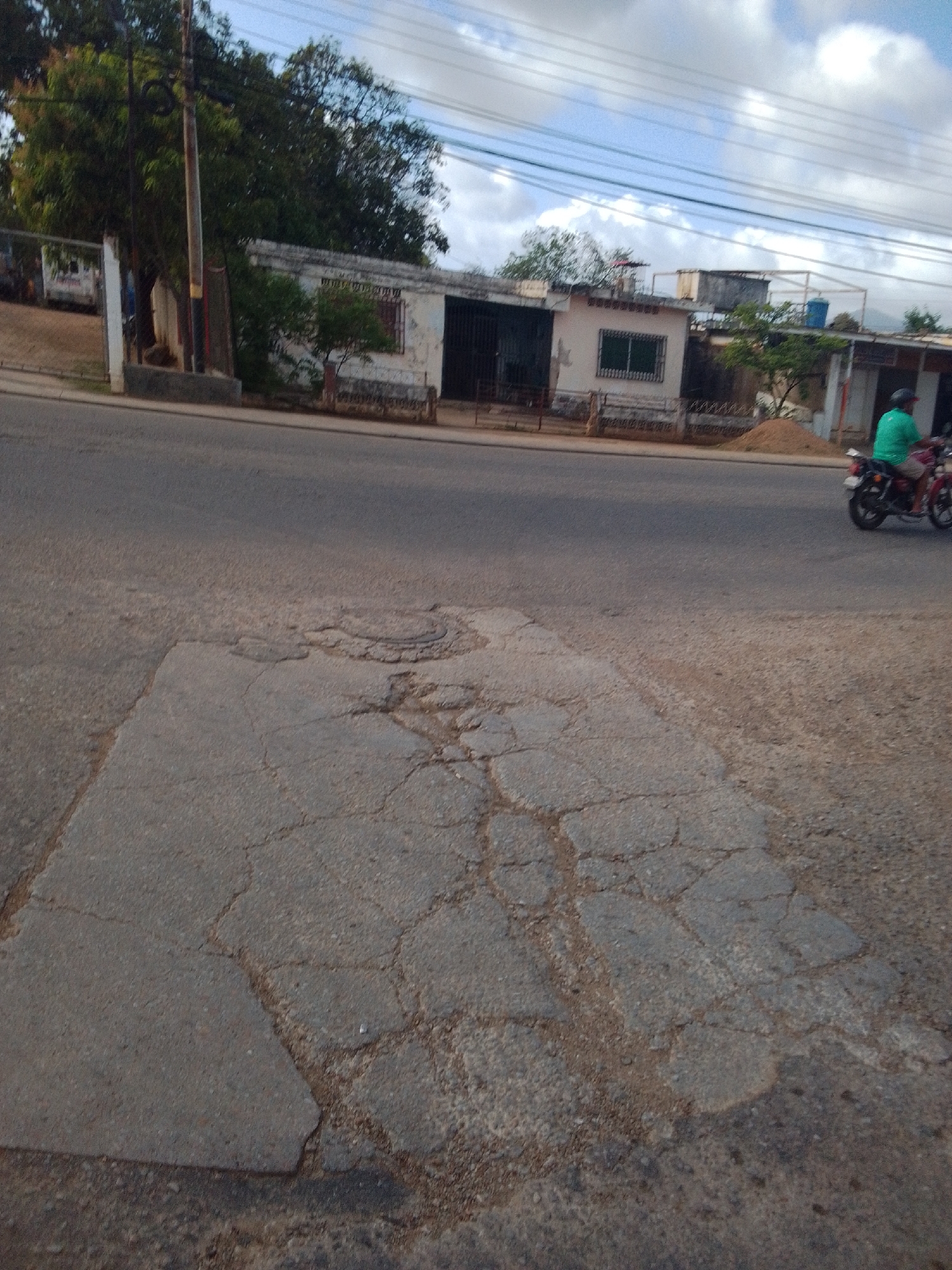 A motorcycle rider wearing a green shirt and a helmet is seen on a cracked asphalt road in Altagracia, Venezuela. The motorcycle is red and black. In the background, there are buildings with white walls and dark windows. Trees and power lines are visible above the buildings. The sky is cloudy. The foreground of the image shows a section of the road with significant cracking and damage.