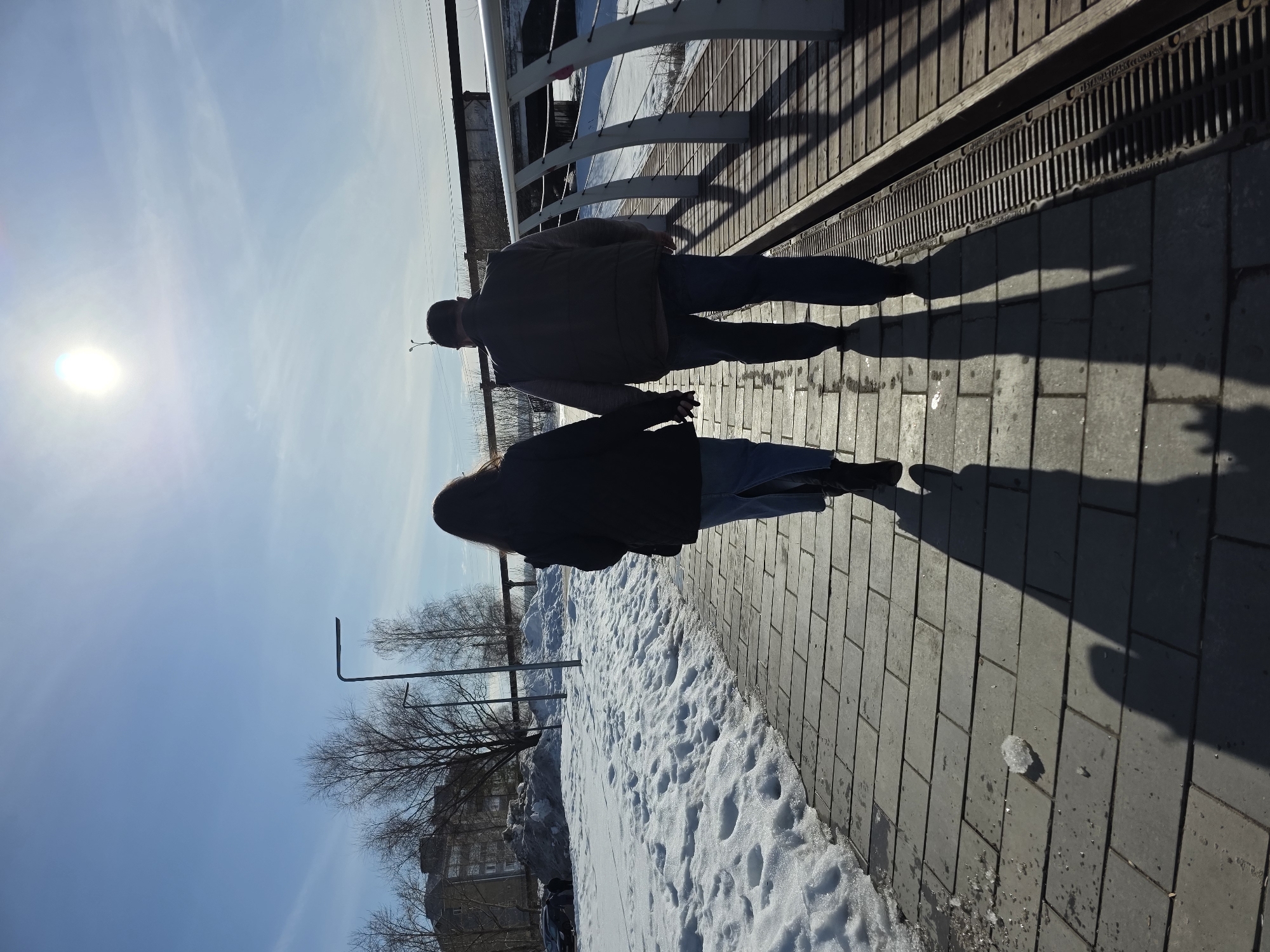 Two adults, a female and a male, are observed from behind, walking hand-in-hand along a paved pathway in Votkinsk, Russia. The female wears a dark jacket, blue jeans, and dark boots. The male wears a dark vest over a lighter shirt, a dark cap, and blue jeans. They cast long shadows forward due to the bright sun positioned high in the upper-center of the clear blue sky.
To the left of the paved pathway, significant accumulations of snow are visible, with some areas showing signs of melting. Beyond the snow, bare deciduous trees and a multi-story building are present. To the right, the paved pathway runs parallel to a wooden boardwalk featuring a continuous metal drainage grate and a white metal railing with curved supports. This railing overlooks a partially snow-covered embankment leading to a body of water. In the background, an elevated structure, possibly a bridge, with visible industrial elements and bare trees, traverses the scene. The overall environment suggests a late winter or early spring day with clear, sunny conditions.