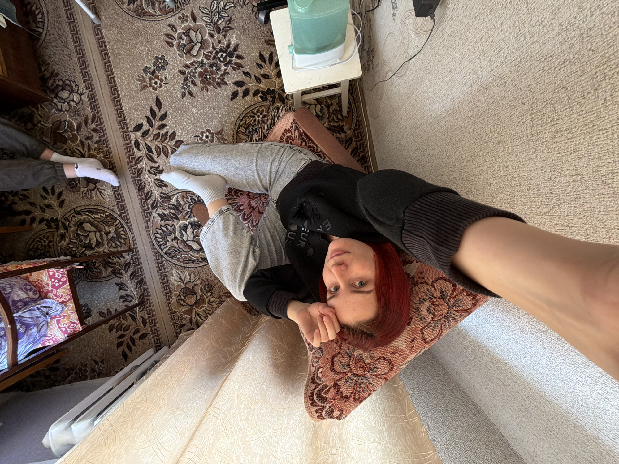 A selfie taken from an elevated angle shows a person with red hair lying in a chair with a red and brown patterned cushion in a room. The person is wearing gray sweatpants and a black sweatshirt. Their left hand is near their face, and their right arm is extended towards the camera. A portion of a second person wearing gray pants and white socks is visible in the lower left corner of the image. The room features a brown patterned rug, light beige wallpaper, and a small white table with a humidifier. Other furniture is also visible, including a wooden chair with a patterned cushion. The location is Chaikovsky, Russia.