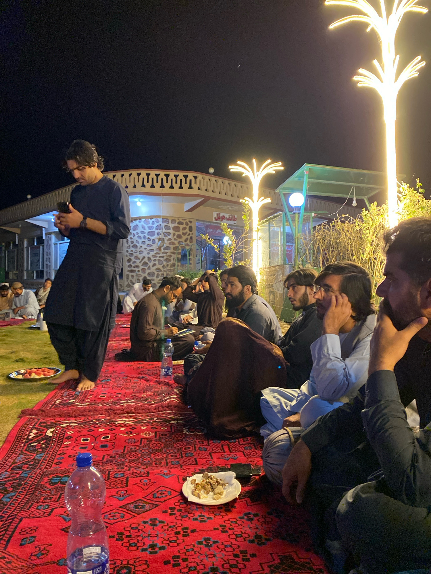 In Gul Kalachah, Afghanistan, a group of men are gathered outdoors at night on a red carpet spread over a grassy area. A large, ornate building with stone accents and decorative railings is visible in the background, illuminated by bright, tree-shaped lights. The men are seated and appear to be engaged in conversation or observing something. Some are holding phones or writing, while others rest their heads on their hands. Food is present on plates, and water bottles are scattered around. The scene suggests a social gathering or a meal shared in an outdoor setting.