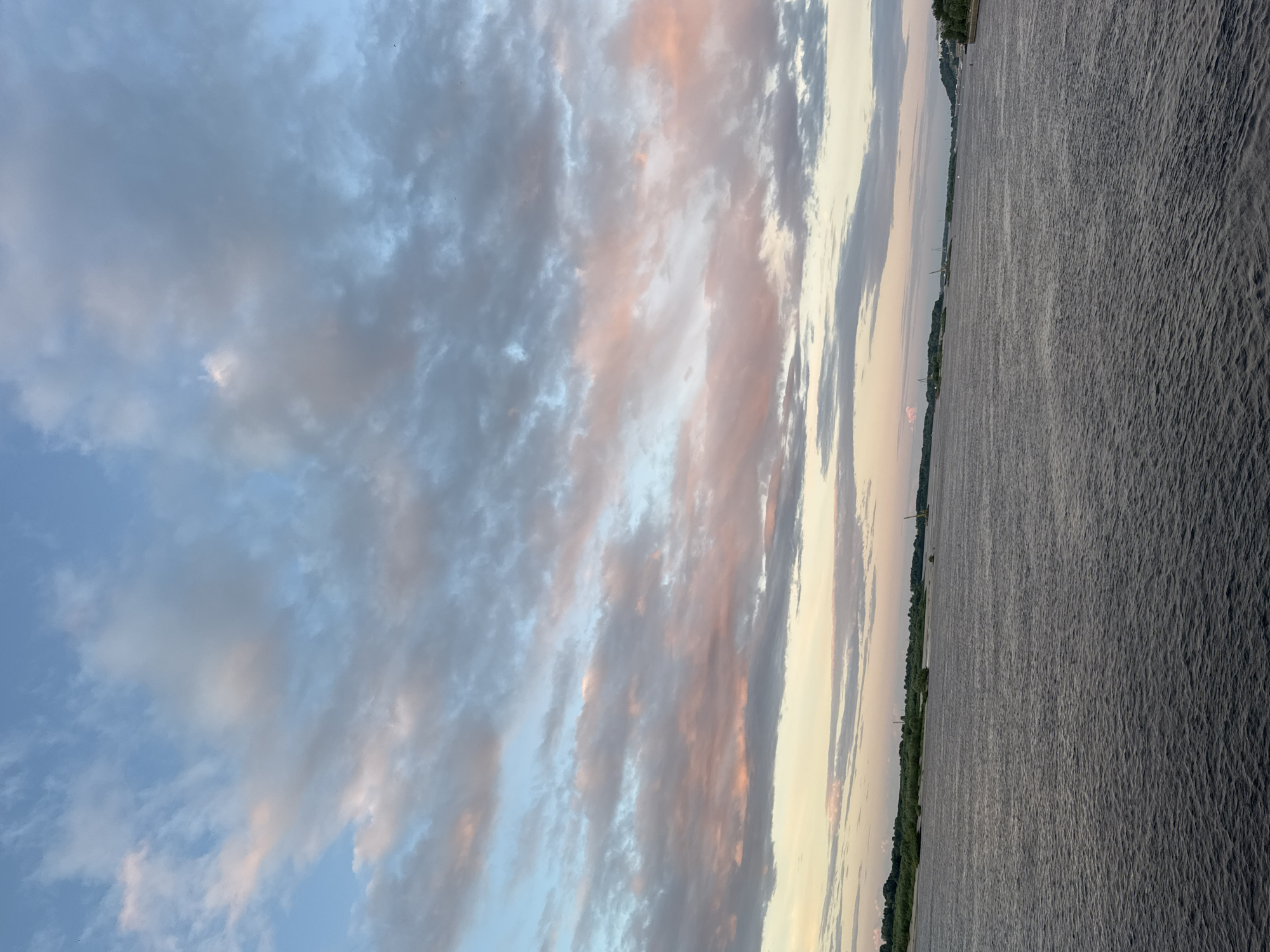 This is a landscape photograph taken at sunset, likely over a body of water such as a river or lake. The image is oriented vertically, with the sky occupying the left two-thirds and the water the right third. The sky is filled with a variety of clouds. Some are wispy and white, while others are denser and appear to be tinged with hues of pink and orange, indicative of sunset. The water is calm with a rippled texture, reflecting the muted colors of the sky.
Along the horizon, a thin strip of land is visible, characterized by lush green trees. In the distance, faint structures that appear to be buildings or towers can be seen along the shoreline, suggesting a developed area. The overall mood of the image is serene and tranquil, capturing the beauty of a natural landscape during the golden hour. The time of day is clearly sunset due to the colors in the sky. The weather appears clear with scattered clouds. There is no visible text in the image.
