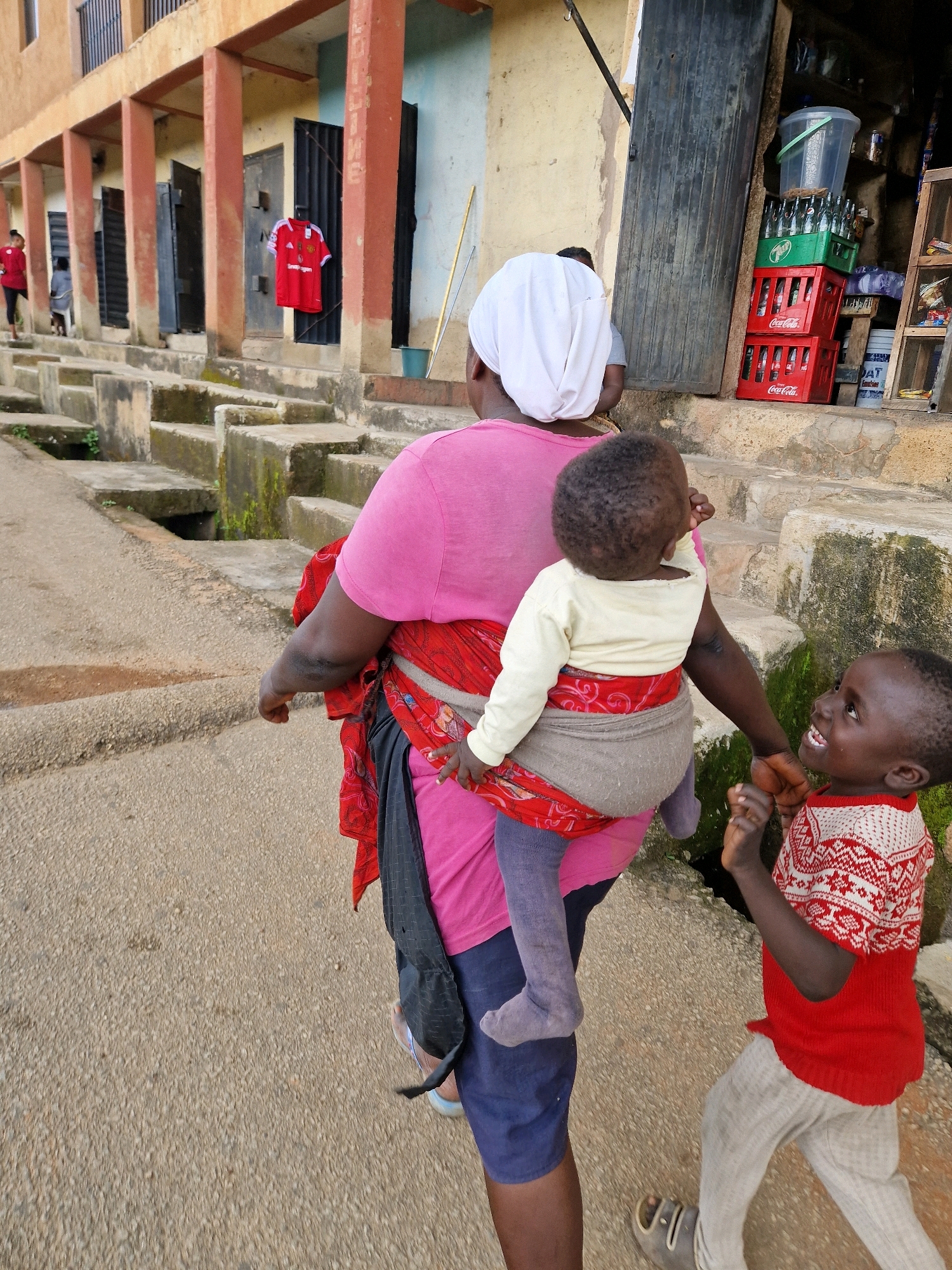 In an outdoor, urban environment of Unknown, Unknown, a woman wearing a pink t-shirt and a white headscarf is seen from behind, carrying a baby on her back. The baby is wrapped in a fabric carrier and wears a light yellow long-sleeved shirt and purple leggings. To the right of the woman, a young child in a red and white patterned sweater and light grey pants smiles and looks up. The ground is a light-colored asphalt surface. In the background, a building with orange-painted pillars and light blue walls is visible, with steps leading up to it. A red t-shirt hangs on a hook in one of the building's openings. To the far right, shelves hold various items, including cases of Coca-Cola bottles and a large plastic container.