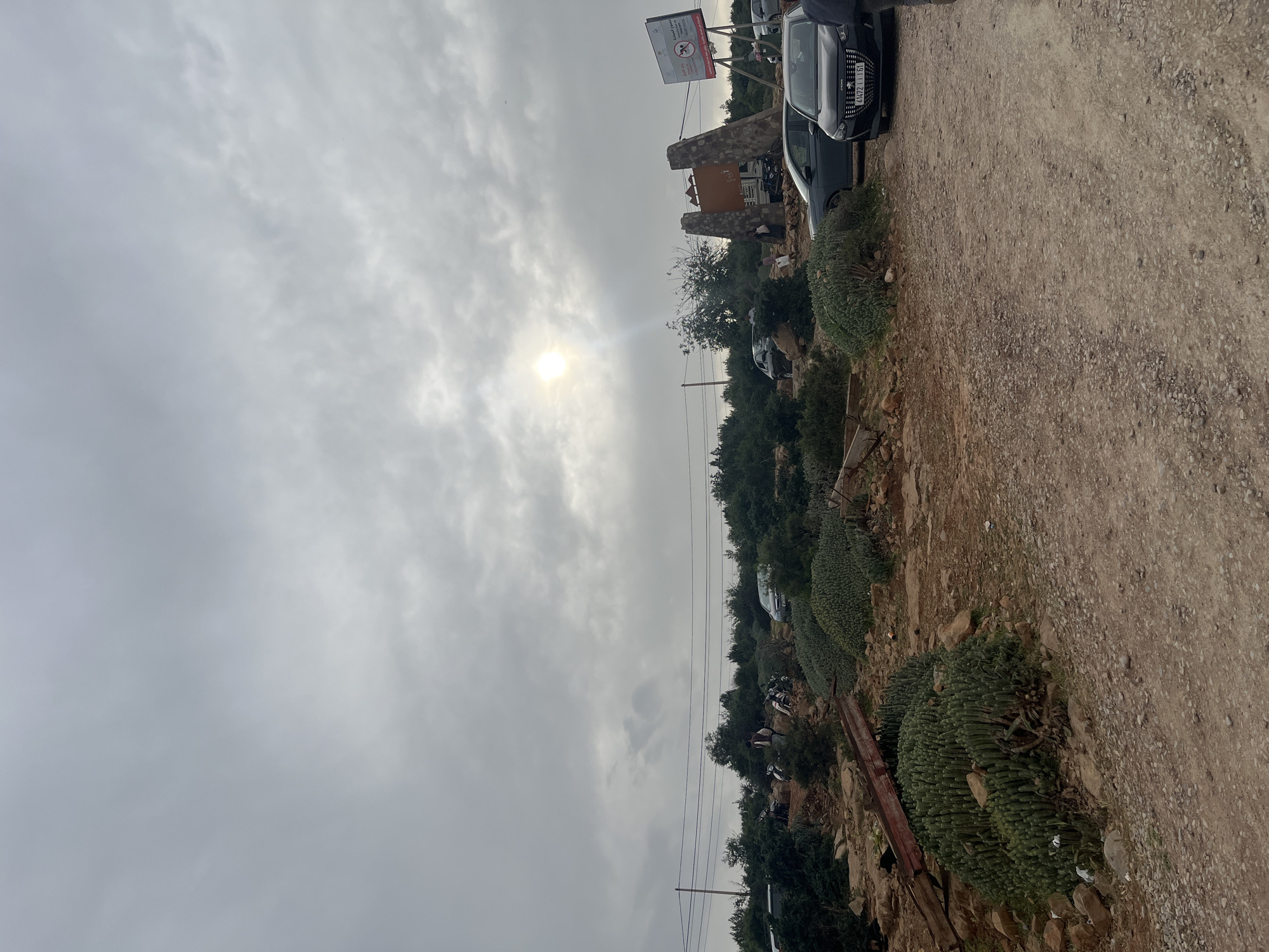 The image depicts an outdoor, sunlit scene under a partially cloudy sky, with the sun prominently breaking through the clouds in the upper left quadrant. The setting appears to be a rural or semi-rural area, possibly a hillside or mountainous region, consistent with Bin El Ouidane, Morocco.
A wide, unpaved dirt road, consisting of light brown soil and small pebbles, stretches along the right side of the frame, leading into the distance. To the left of the road, a rugged, rocky terrain is covered with lush, green vegetation, including distinctive columnar succulents and various trees and bushes.
In the mid-ground, several vehicles are parked. A dark-colored car, possibly a Peugeot, is clearly visible in the foreground with the Moroccan license plate "A4721151." Further down the road, more cars are parked, indicating a stopping point or a parking area. Near the cars and vegetation, two rustic stone pillars flank a wooden sign, forming what appears to be an entrance. Above this structure, a white sign with Arabic text displays "مرحبا بكم" (Marhaba Bikom), meaning "Welcome," along with a logo of a person swimming and other unreadable text, suggesting a tourist or recreational establishment like a cafe or camping site. Faint power lines and poles are also visible across the landscape. A few people are indistinctly seen near the parked vehicles and structures.
The time of day is clearly daytime, with the sun actively illuminating the scene through the clouds. The weather is overcast but bright, creating a diffused light. The overall atmosphere suggests an active, yet potentially tranquil, outdoor destination.
