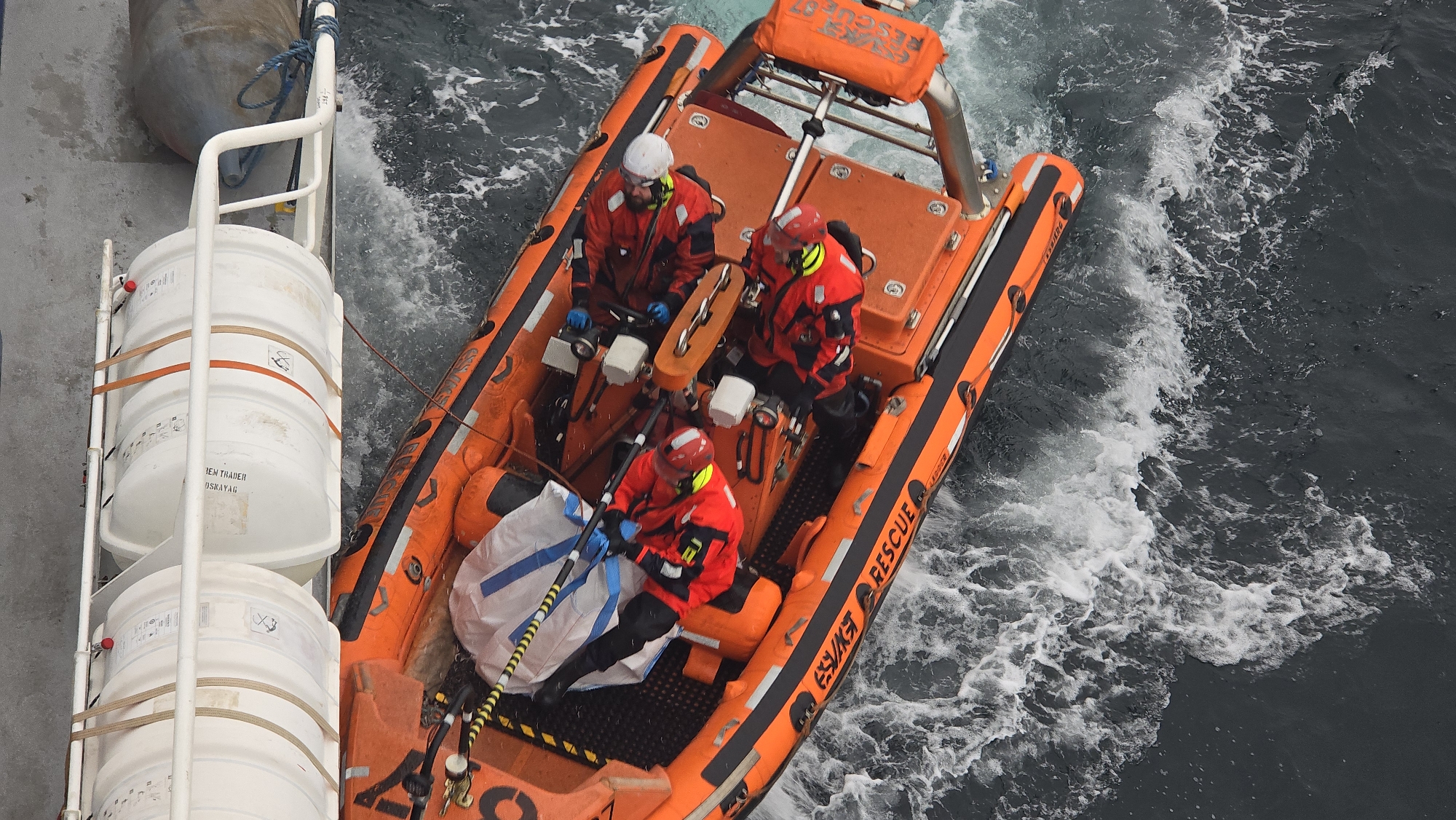 An eye-level, medium-shot photograph shows an orange rescue boat, likely a Rigid Hull Inflatable Boat (RHIB), in choppy water. Three people in bright red and black suits are aboard the boat. The person in the bow of the boat is kneeling and holding what appears to be a long, white and blue bag with black and yellow stripes. Two other people are at the helm, wearing white and red helmets. The side of the boat has the word "RESCUE" printed on it. Adjacent to the boat, on the left side of the image, is a white structure with two large, round, white canisters. The water around the boat is dark with white foam. The location is Unknown, Unknown.