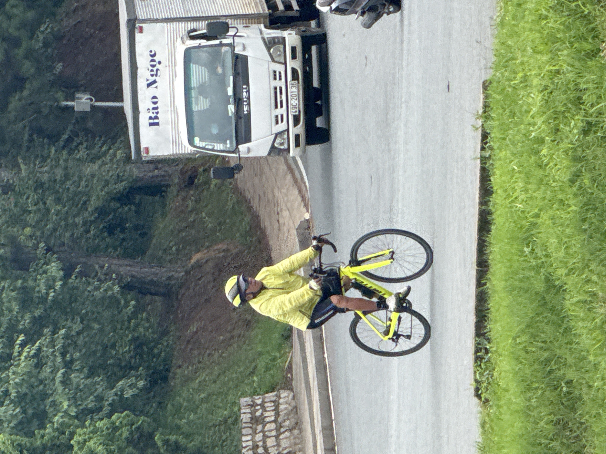 An image, oriented upright for viewing, depicts a male cyclist riding a bright yellow road bike on a paved road. The cyclist wears a matching yellow long-sleeved jersey, black shorts, a yellow helmet, and sunglasses, appearing focused on their activity.
The road is bordered by lush green vegetation, including trees and dense grass, suggesting a natural or park-like environment. A low stone wall runs alongside the left edge of the road. Further down the road in the background, a white box truck is visible. The truck has "Bảo Ngọc" written in blue Vietnamese script on its side panel and "ISUZU" on its front grille. Its license plate displays "49C-201.36". A utility pole with a small attached box, possibly a camera or sensor, can also be seen in the left background.
The scene takes place during the daytime, under what appears to be an overcast or hazy sky, providing soft, diffused lighting with no harsh shadows. Based on the visible Vietnamese text and license plate format, the location is confirmed to be in Vietnam, consistent with the provided context of Phường Xuân Hương, Đà Lạt.