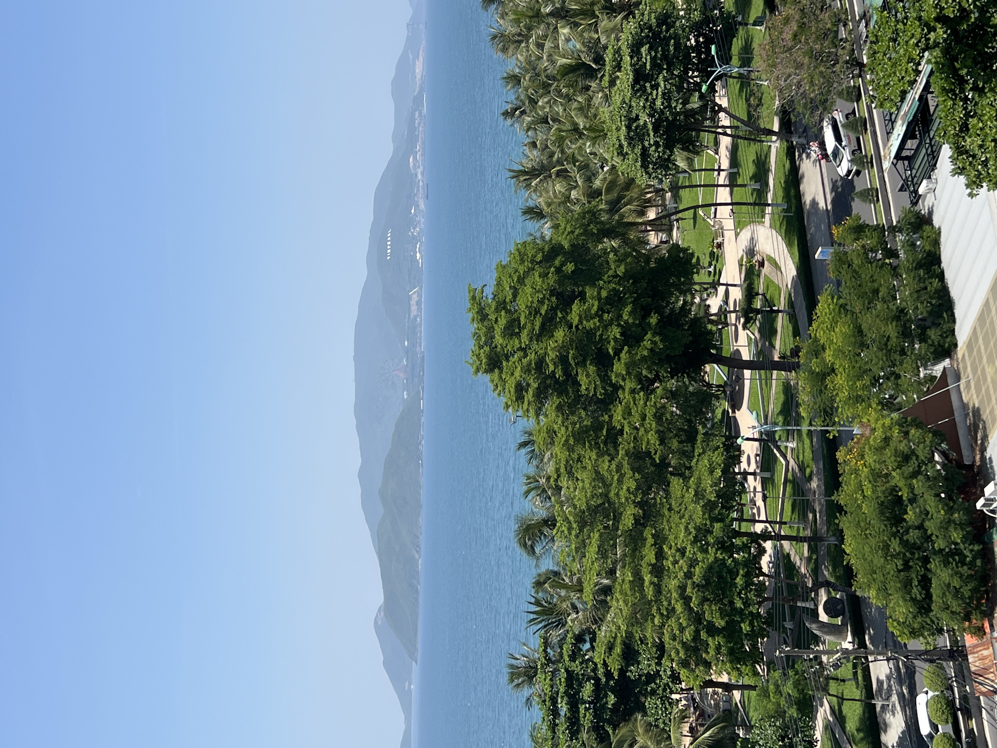 This is a vertical, high-angle shot of a coastal scene in Nha Trang, Vietnam. The image captures a clear blue sky above a calm, blue ocean. In the distance, a lush green mountain range is visible, with "VINPEARL" written in large white letters on the side of one of the mountains. In the mid-ground, a park with well-maintained green lawns, winding pathways, and scattered trees and palm trees is situated between the ocean and a street. There are no people clearly visible in the park. On the street in the foreground, a white SUV and a motorcycle are present. The overall impression is a bright, sunny day in a tropical coastal city.
