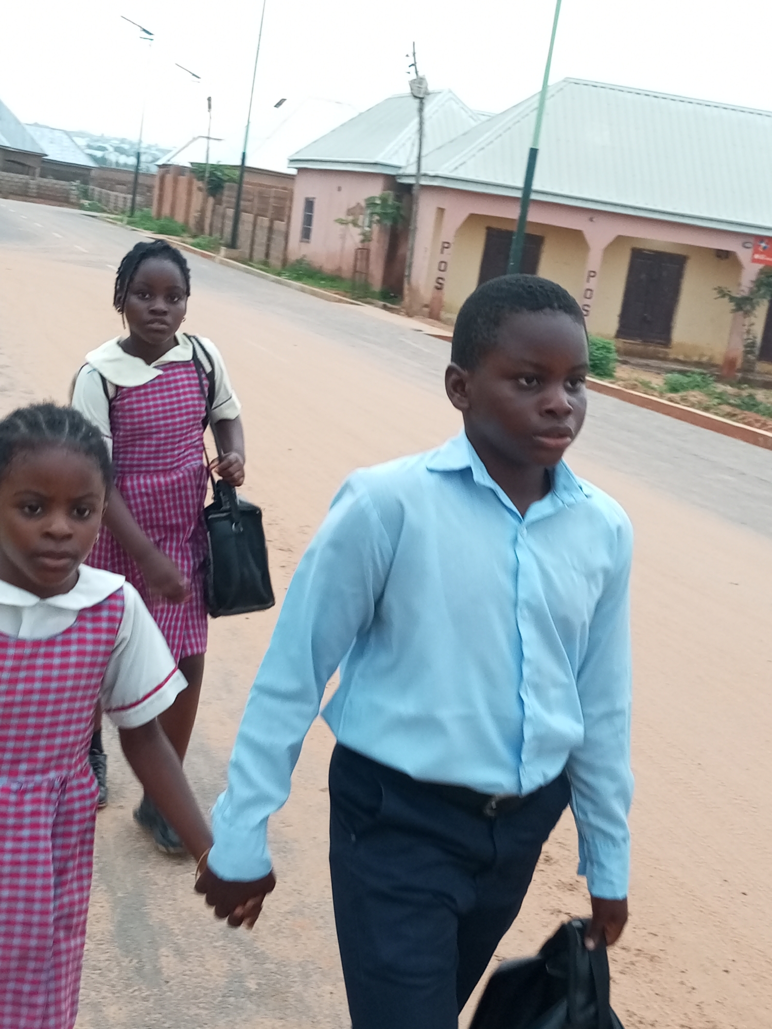 In Tempure, Nigeria, three children are walking on a dusty road. A boy in a light blue button-down shirt and dark pants leads the way, carrying a black bag in his right hand. He holds the left hand of a girl walking beside him. This girl wears a pink and white checkered dress and carries a black bag in her right hand. Another girl, closer to the camera, also wears a pink and white checkered dress and looks towards the viewer. In the background, buildings with light-colored roofs and walls line the street. The sky is overcast.