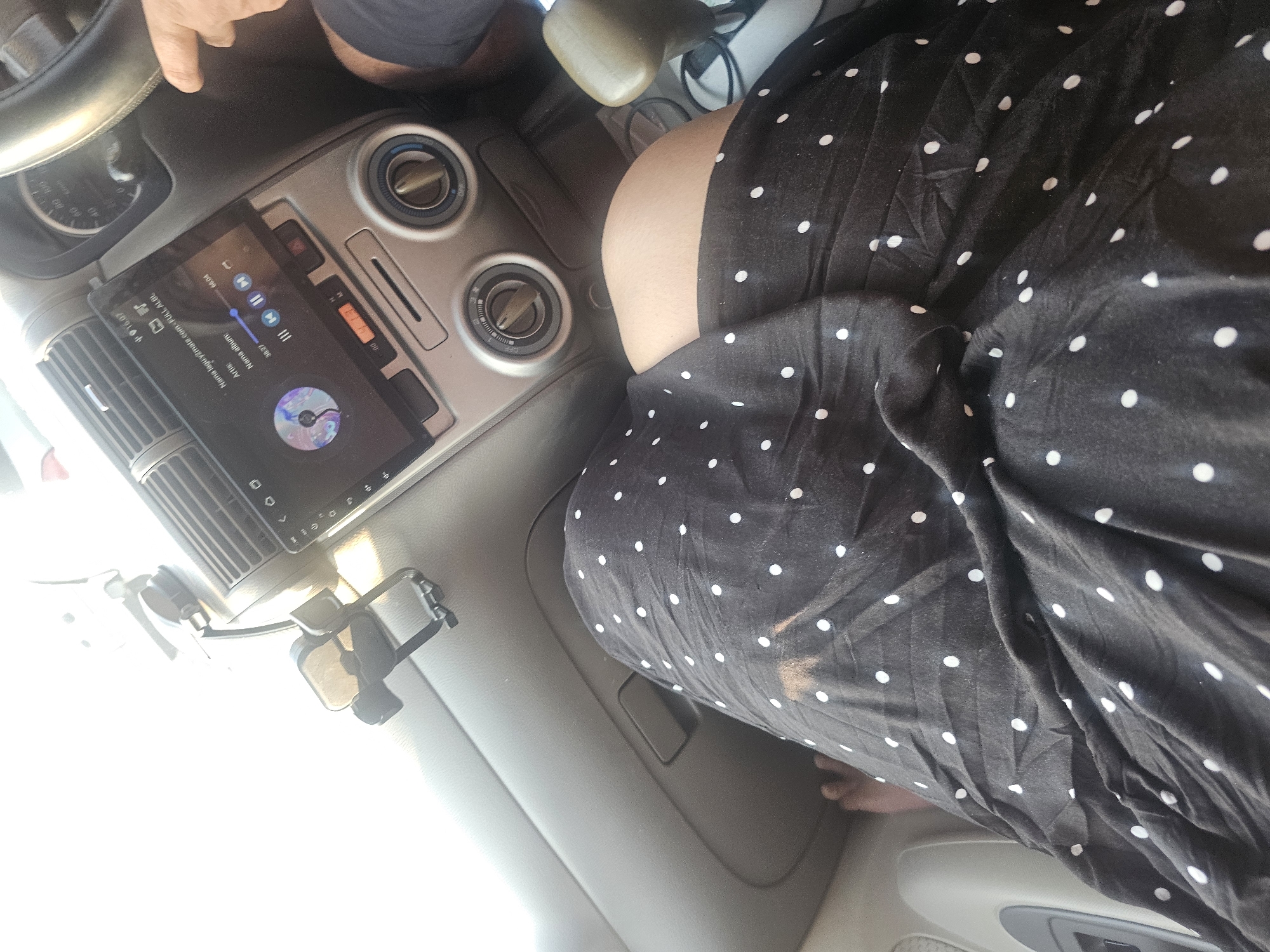 A person's legs, draped in a black, polka-dotted skirt, are positioned in the passenger seat of a vehicle. The dashboard is visible, featuring a car stereo with a screen displaying music information, a climate control panel with rotary knobs, and air vents. A phone holder is attached to the dashboard. The scene is illuminated by natural light, suggesting it is daytime. The location is identified as Semanding, Indonesia.