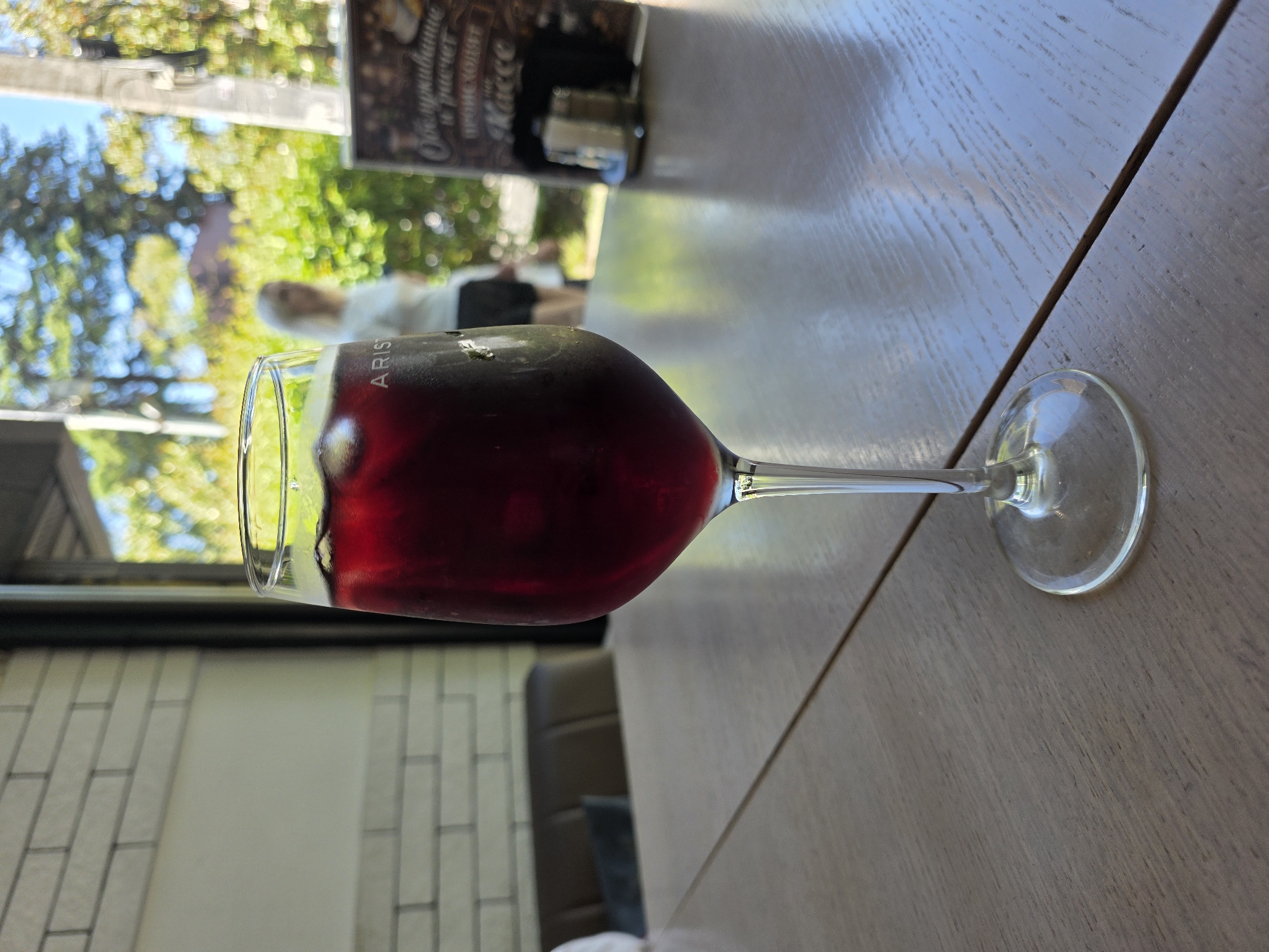 A close-up, eye-level shot features a tall glass filled with a dark red beverage, likely wine or sangria, on a light wooden table. Ice cubes are visible within the drink. The glass has "ARISTO" printed on it. In the background, a blurred figure of a woman with blonde hair wearing a white top stands near lush green foliage and trees. To the right of the woman, a blurred menu or sign with Cyrillic text is partially visible, along with salt and pepper shakers. The setting appears to be an outdoor seating area of a cafe or restaurant, possibly in Sochi, Russia, given the presence of Cyrillic text and the warm, sunny outdoor environment with trees. The table is made of light-colored wood with a visible seam down the middle.