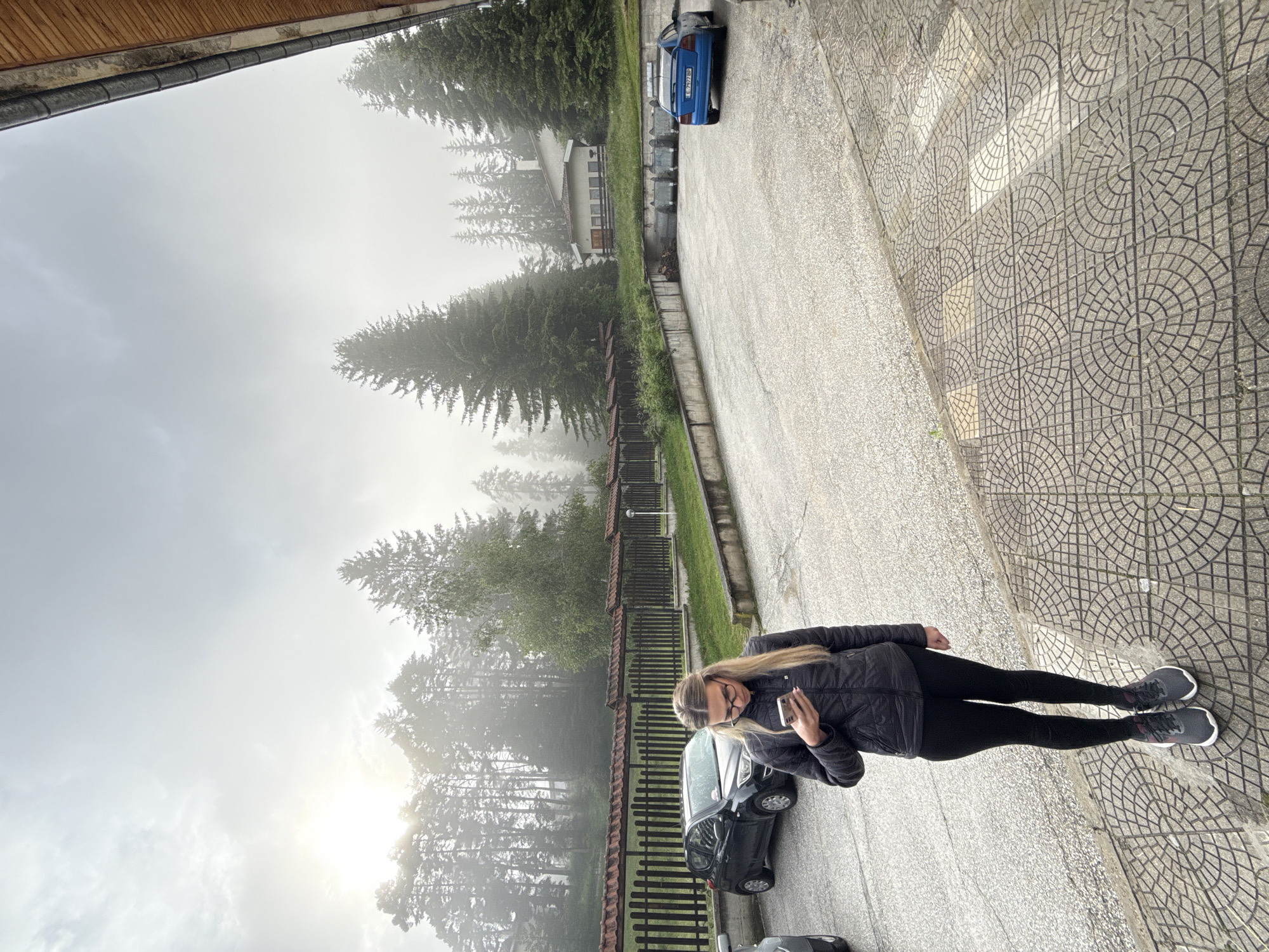 A young woman with blonde hair and glasses stands on a patterned tile walkway, looking at her phone. She is wearing a black puffer jacket, black leggings, and grey athletic shoes. Behind her is a paved parking lot with a black car parked on the left and a blue car parked on the right. In the background, a wooden fence with red roofing runs along the edge of a grassy area. Beyond the fence, a dense forest of tall pine trees rises up a hill, with a building partially visible amongst them. The sky is overcast and hazy, with the sun faintly visible through the clouds, suggesting a misty or foggy morning. The location is identified as Yundola, Bulgaria.