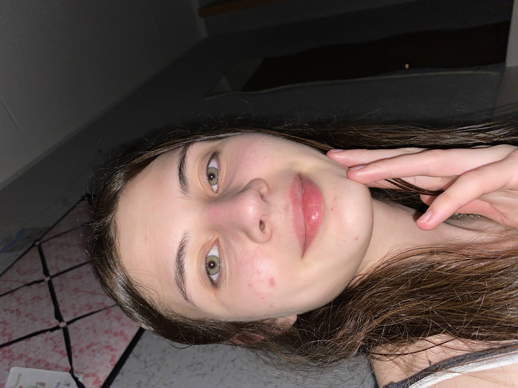 A young woman with light skin, brown hair, and green eyes poses for a selfie. Her face is angled towards the camera, with her right hand touching her cheek. She has some blemishes on her face. The background appears to be a dimly lit room, with a patterned surface visible in the lower left corner. There is no indication of the city of Votkinsk, Russia.