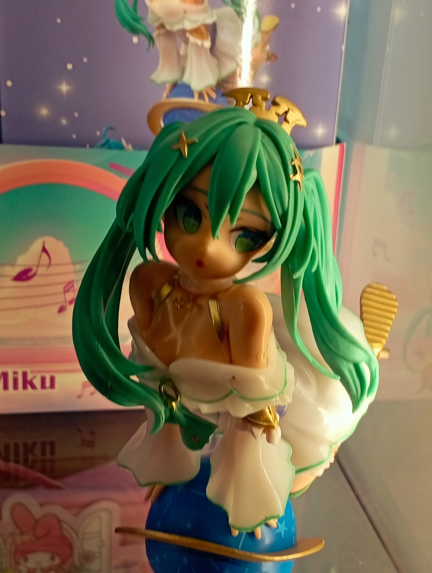 The image displays a Hatsune Miku figurine positioned centrally in an indoor setting. The figurine features long, teal-green twin-tails, large green eyes, and a light-toned complexion. She wears a white, flowing dress with subtle green trim and gold accents, including a gold crown-like headpiece with star motifs and additional gold star ornaments in her hair. Her upper chest and shoulders are bare, adorned with a gold necklace-like detail. The figurine stands on a blue sphere, stylized as a planet, with white stars and a gold ring around its equator.
In the background, multiple pieces of packaging are visible. Directly behind the figurine, a purple box displays various white stars, some appearing as four-pointed starbursts. Above this box, a portion of another figurine, possibly the same character in a white dress, is visible. Below the purple box, a light-colored box features a rainbow arc composed of pink, orange, yellow, green, and blue stripes. This box also displays purple musical notes on staff lines and the word "Miku" in purple text. Below this, a third box is partially visible, showing a pink background with a cartoon character resembling My Melody, a white rabbit with a pink hood, depicted inside a window frame. The partial word "Nikn" is also visible on this lowest box. The lighting illuminates the figurine from the front, creating soft highlights and shadows. The entire scene suggests a display setup, possibly on a shelf or in a retail environment. No elements within the image visually identify a specific geographic location, including the city of Porlamar, Venezuela.