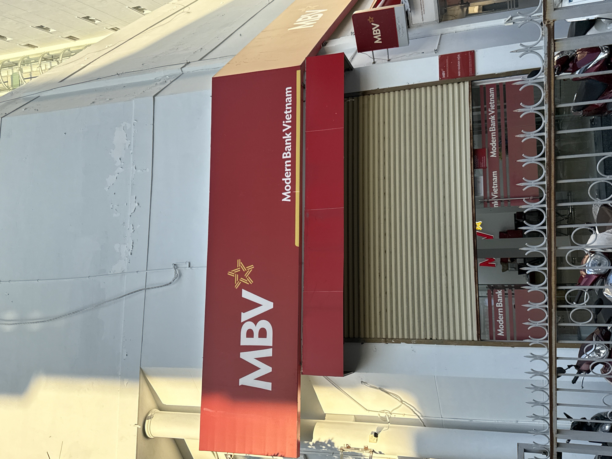 The image shows the exterior of a Modern Bank Vietnam branch in Nha Trang, Vietnam. The bank has a prominent red sign with "MBV" in white lettering and a stylized yellow star. Below this, another red awning displays "Modern Bank Vietnam". A closed metal security shutter covers the entrance. Through a small window, the interior of the bank is partially visible, with more red branding and what appear to be desks or counters. A white metal fence with circular and pointed decorative elements runs in front of the bank. Two motorcycles are parked behind the fence, suggesting a street-level location in an urban area. The building facade is white and shows some signs of weathering and peeling paint. The lighting suggests it is either early morning or late afternoon, with a strong shadow cast on the upper part of the building.